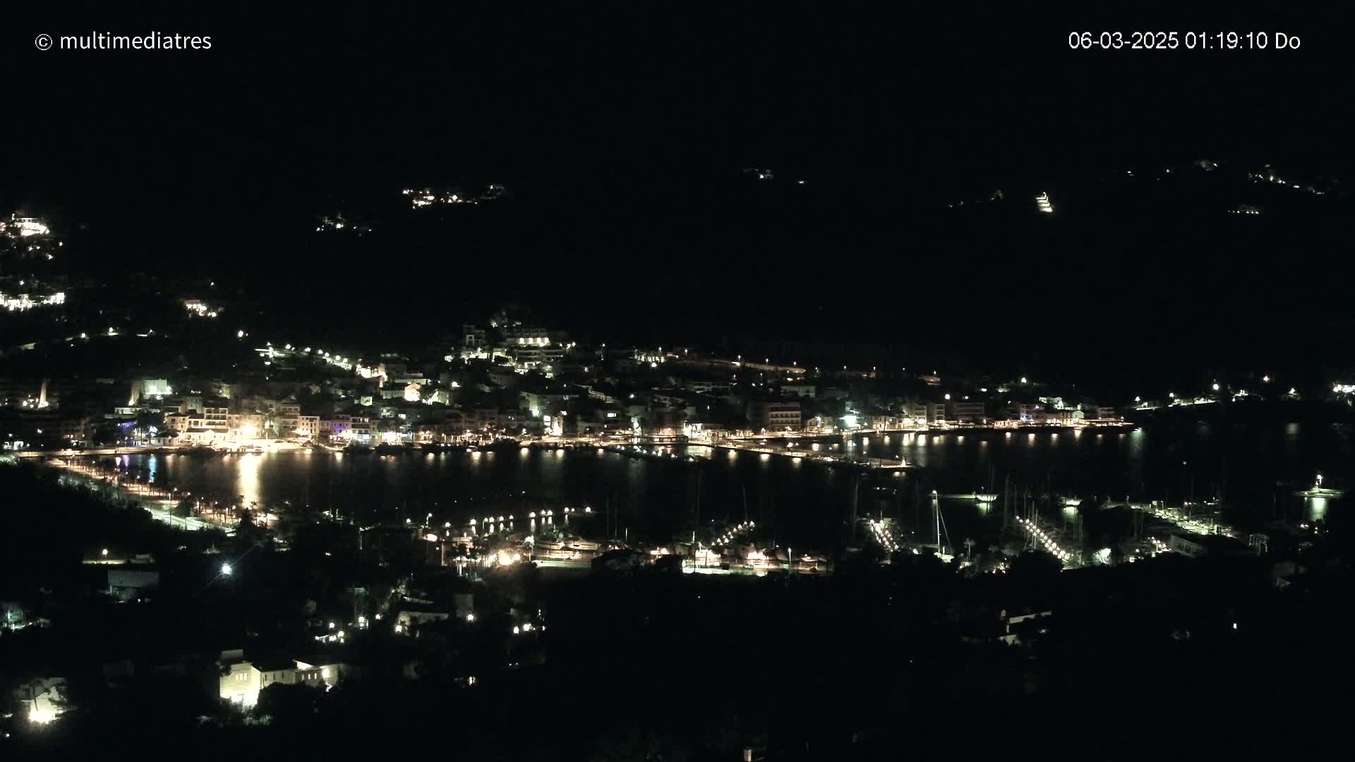 A nighttime view of a harbor town with its lights illuminating the waterfront and surrounding hills under a dark sky.
