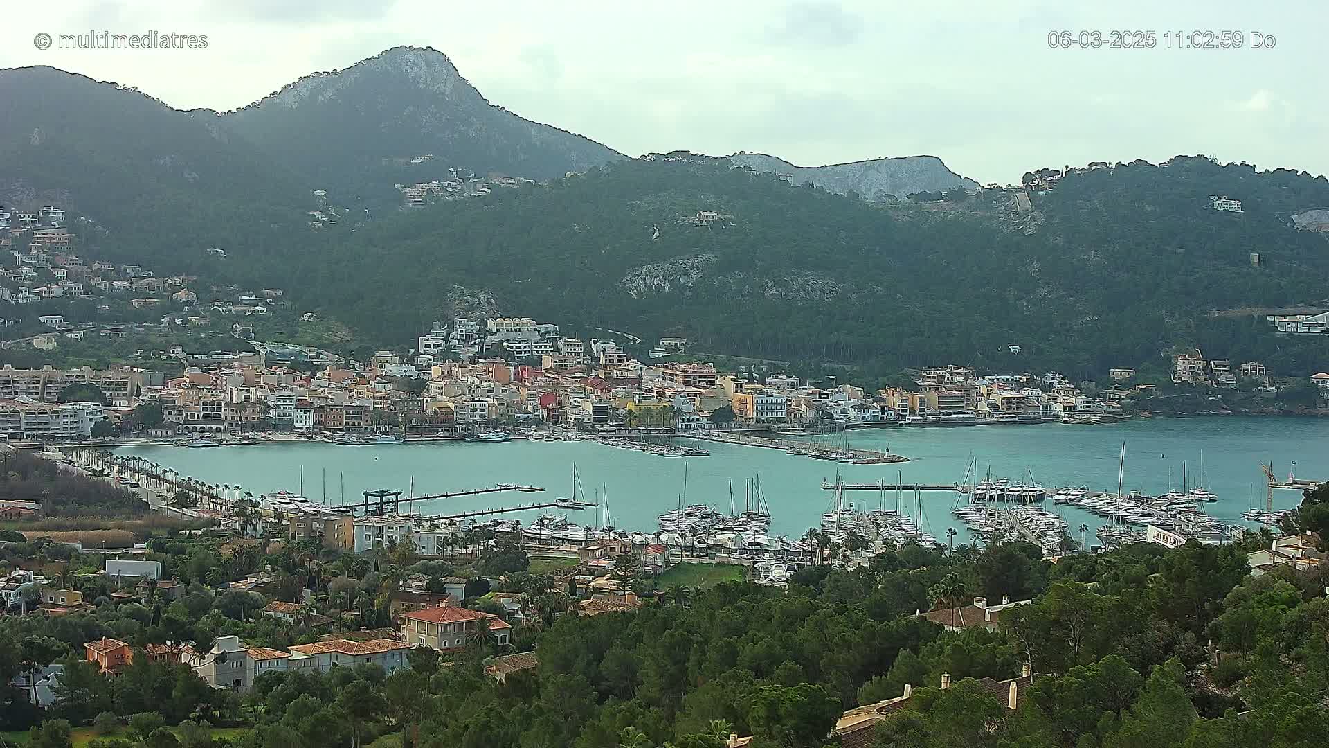 A coastal town with a marina filled with boats sits nestled at the base of a mountain range under an overcast sky.