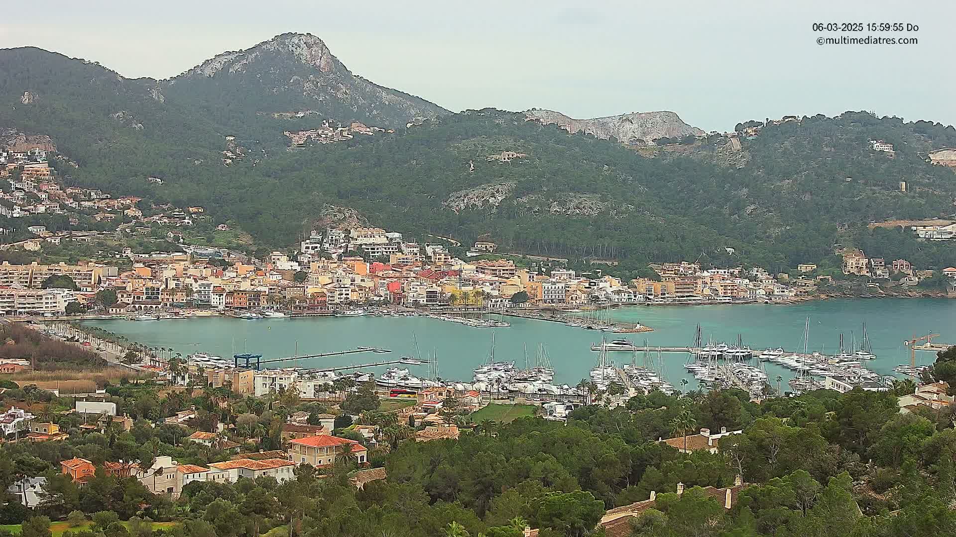A coastal town nestled in a bay, surrounded by green hills and mountains under an overcast sky, features a marina filled with numerous boats.