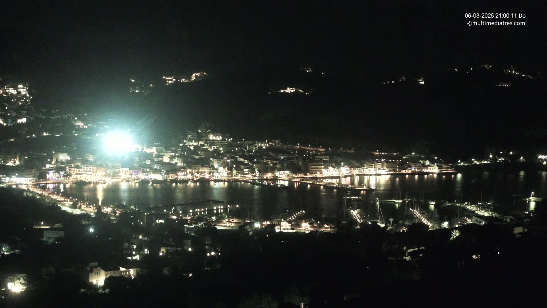 A nighttime, high-angle view shows a brightly lit coastal town nestled between dark hills, with a calm harbor filled with boats.