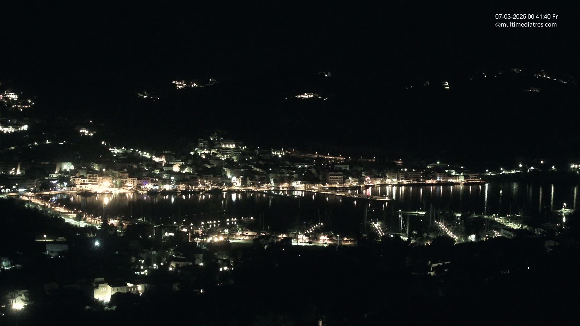 A nighttime, aerial view of a coastal town illuminated with lights, nestled beside a calm body of water reflecting the city lights.