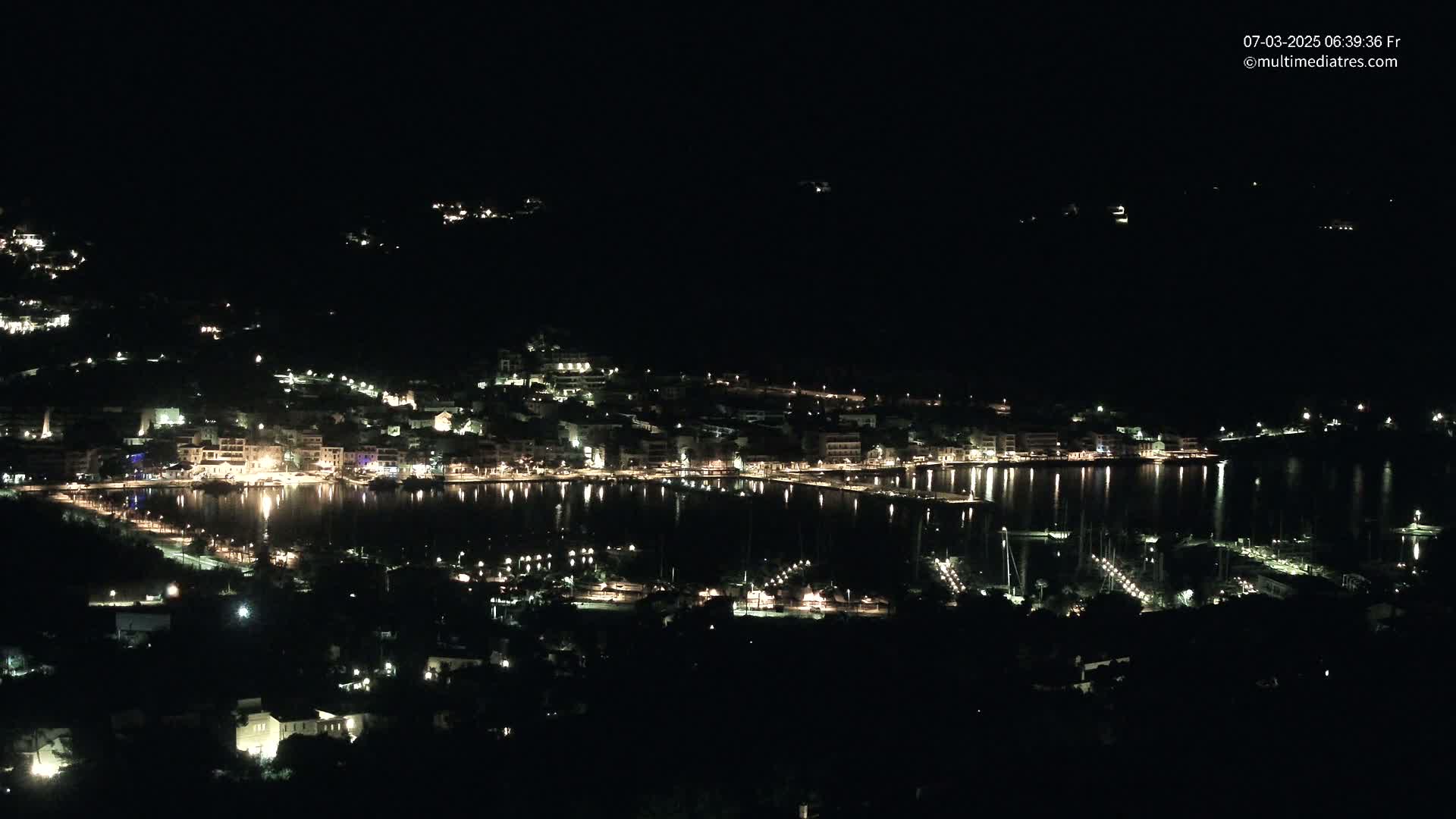 A nighttime view of a coastal town illuminated by lights, with calm water reflecting the lights, and surrounding dark hills.
