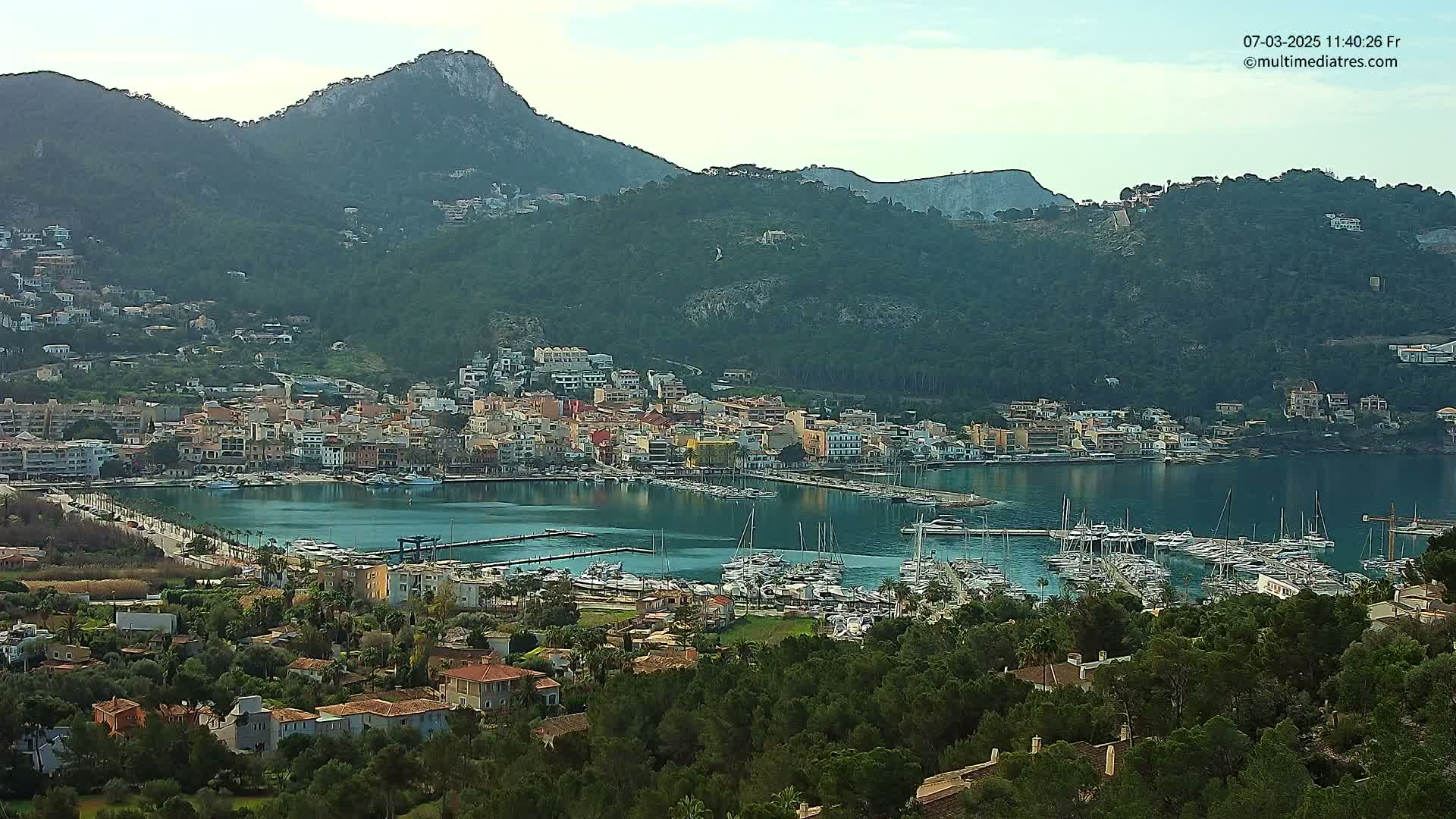 A coastal town nestled in a mountainous region, with a calm harbor filled with numerous boats, under a mostly clear sky.