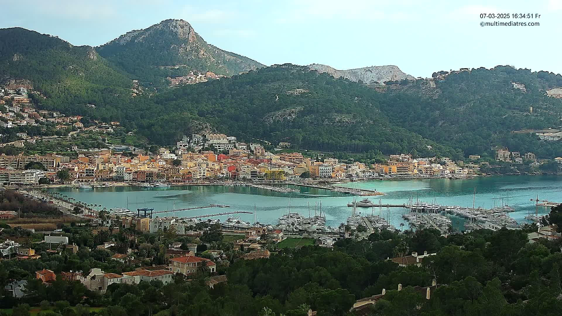 A coastal town with a marina full of boats is nestled in a valley between green, mountainous hills under a mostly clear sky.