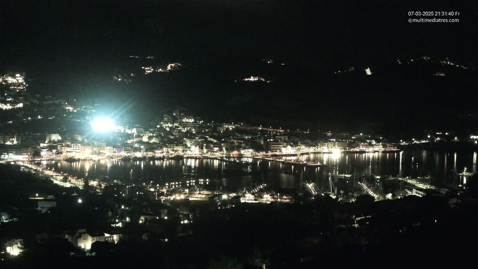 A nighttime, high-angle view shows a coastal town illuminated with lights along a calm body of water, surrounded by dark hills.