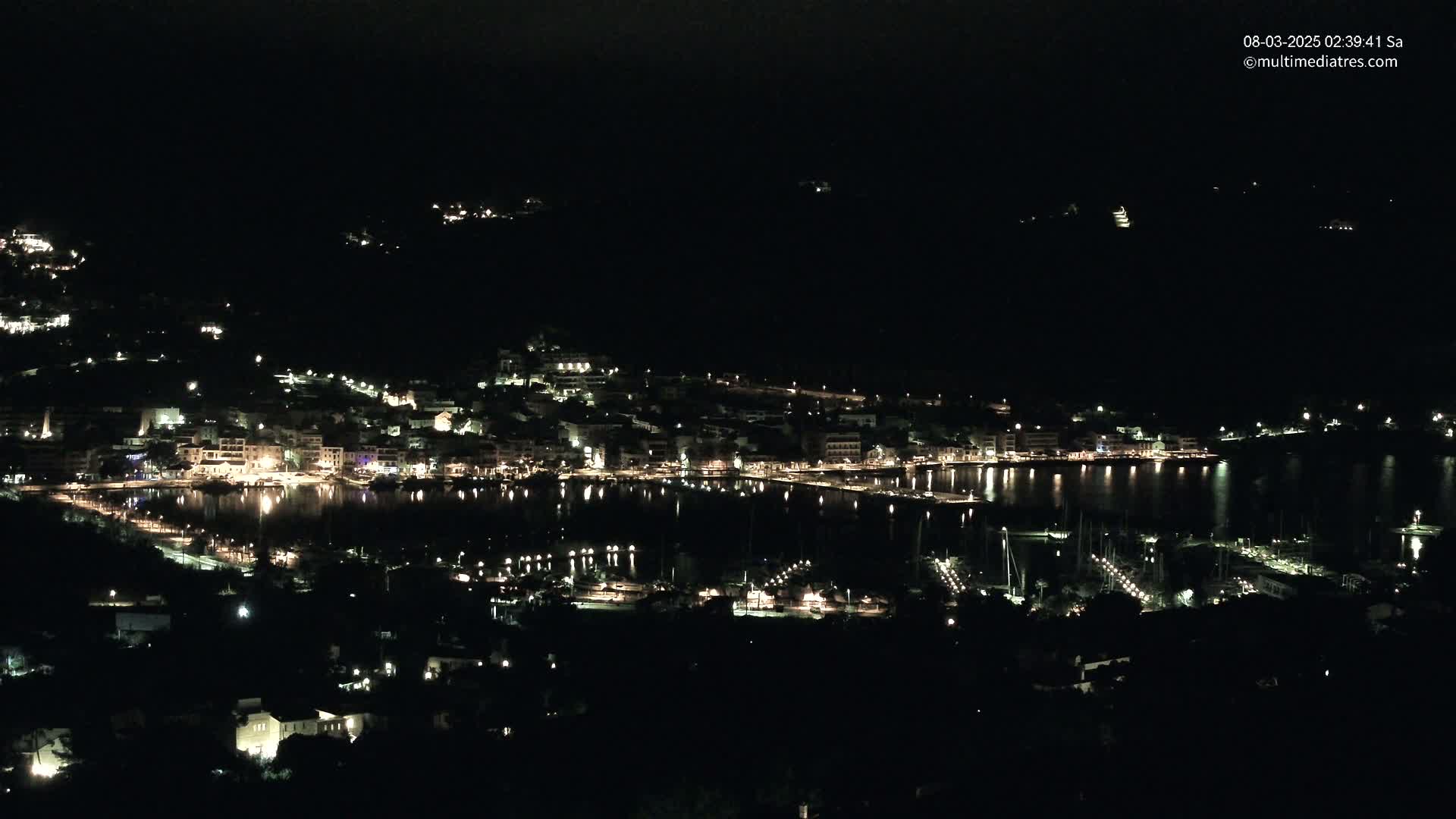 A nighttime, aerial view of a coastal town illuminated by many lights, with a dark, calm body of water reflecting the lights.