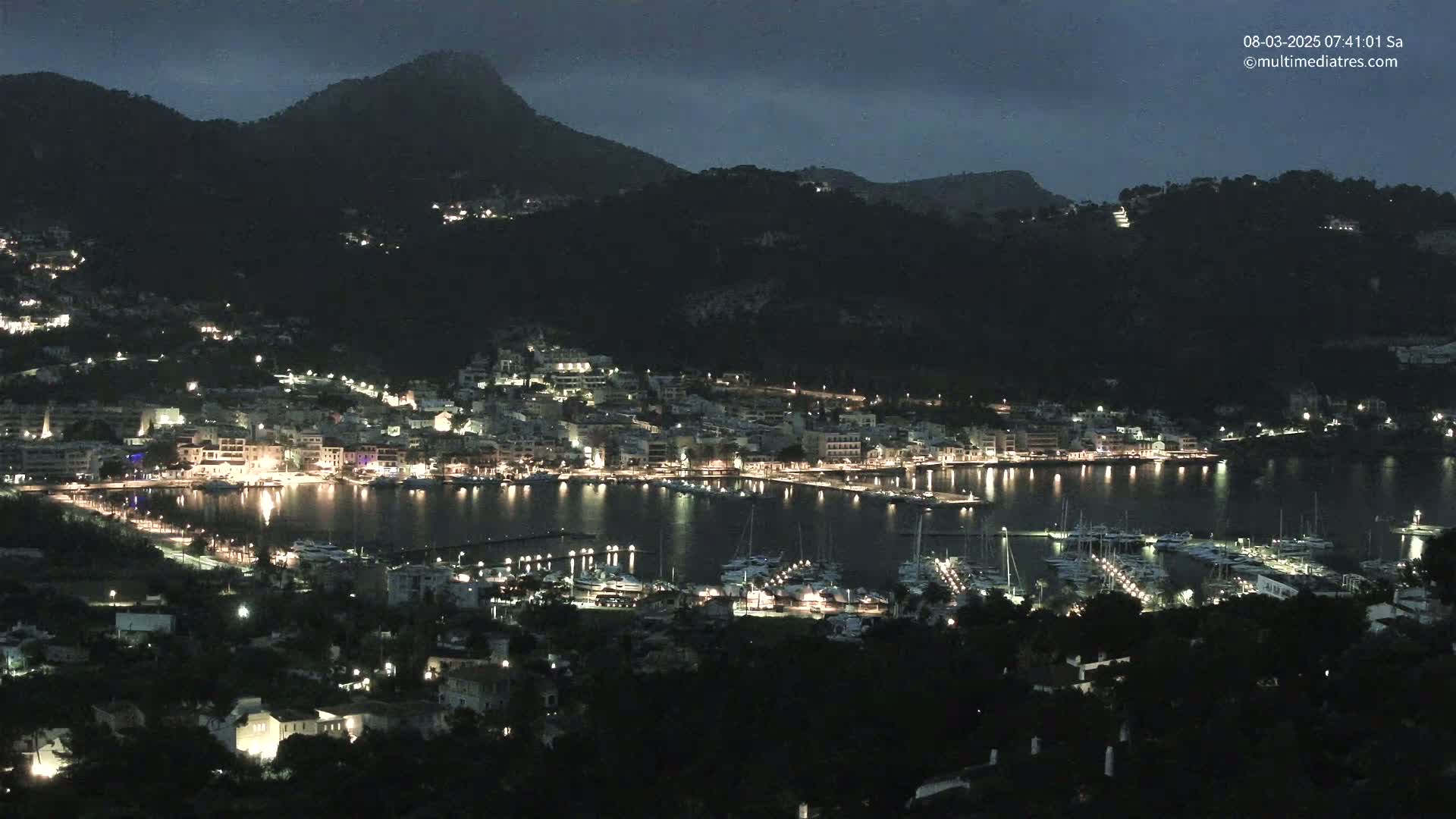 A nighttime view of a harbor town nestled between dark hills under a cloudy sky, with many boats docked in the harbor and lights illuminating the buildings and waterfront.