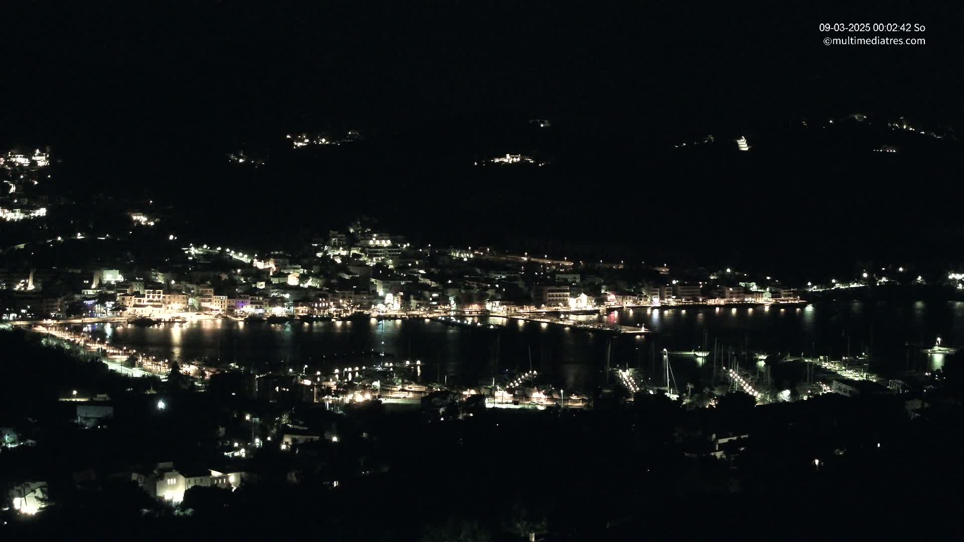 A nighttime, aerial view of a coastal town with many lights illuminating buildings and boats in a harbor.