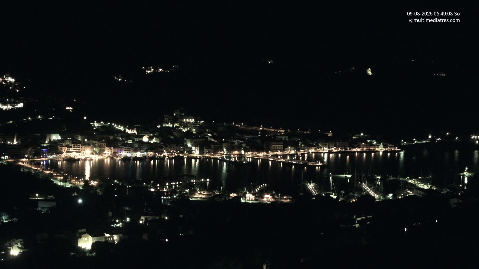 A nighttime, aerial view of a coastal town illuminated by lights, with a calm body of water reflecting the lights.