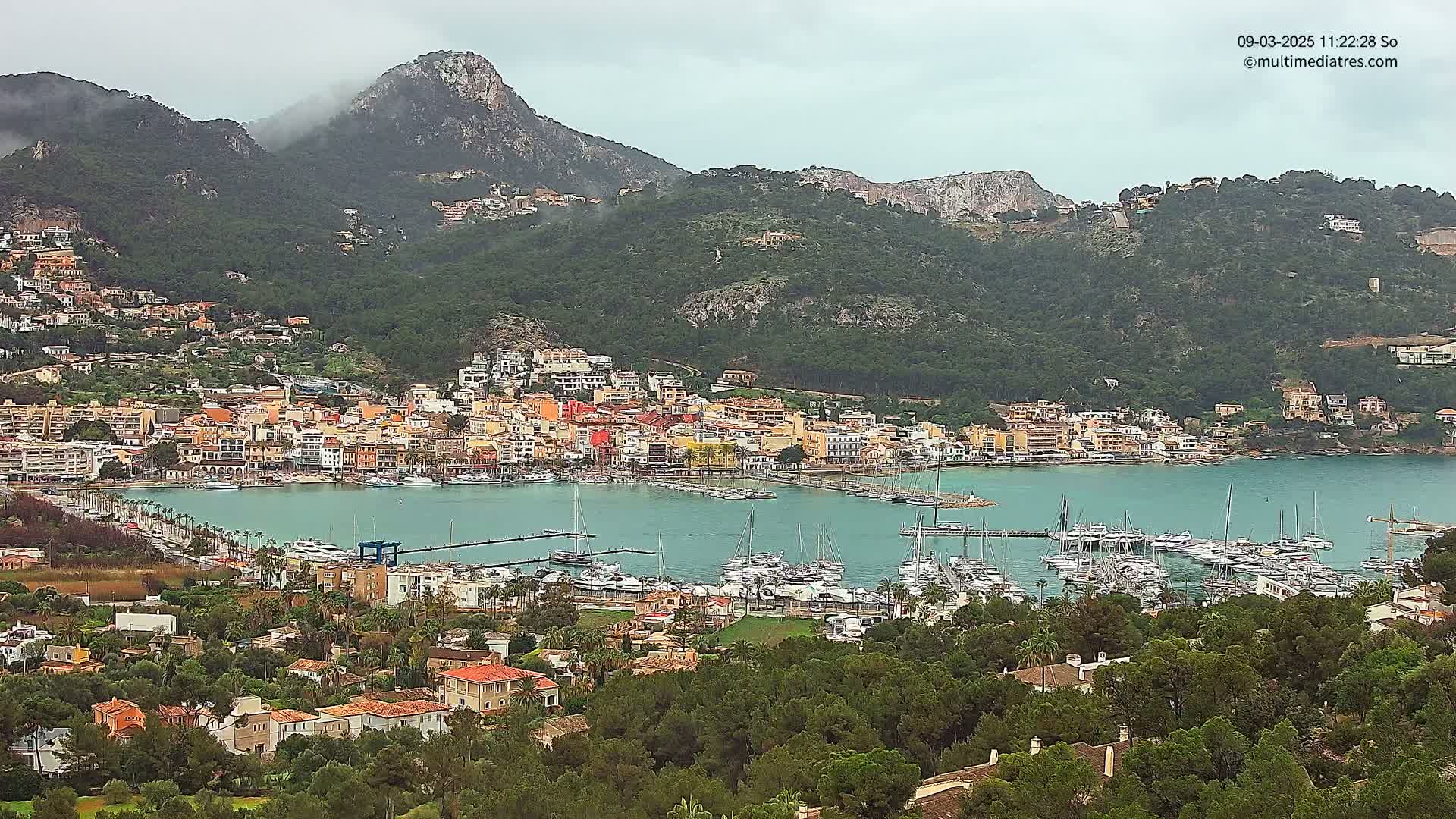 A coastal town nestled between green hills under an overcast sky, with a harbor full of sailboats.