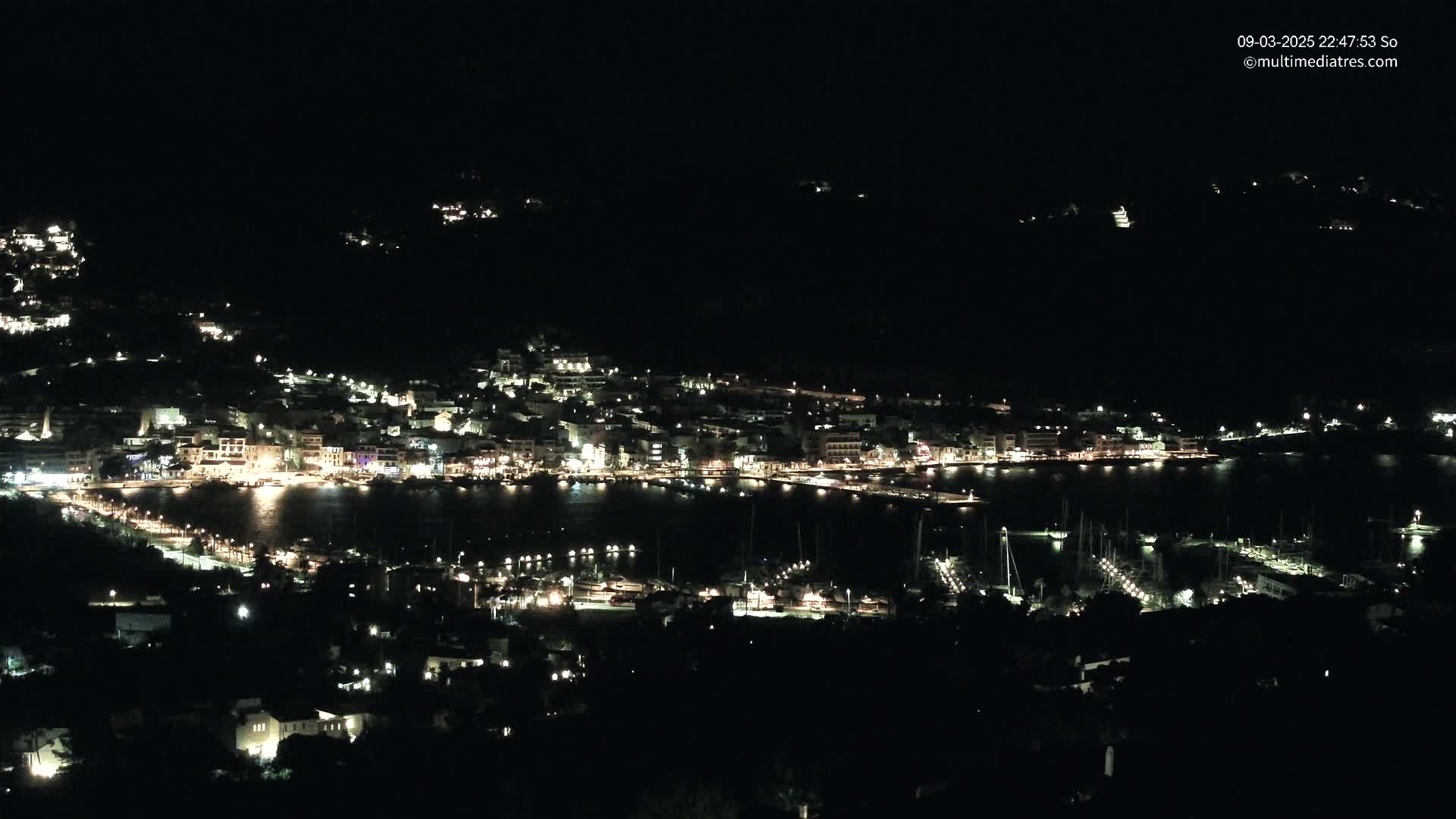 A coastal town at night, illuminated by many lights, is nestled against dark hillsides beside a harbor filled with boats.