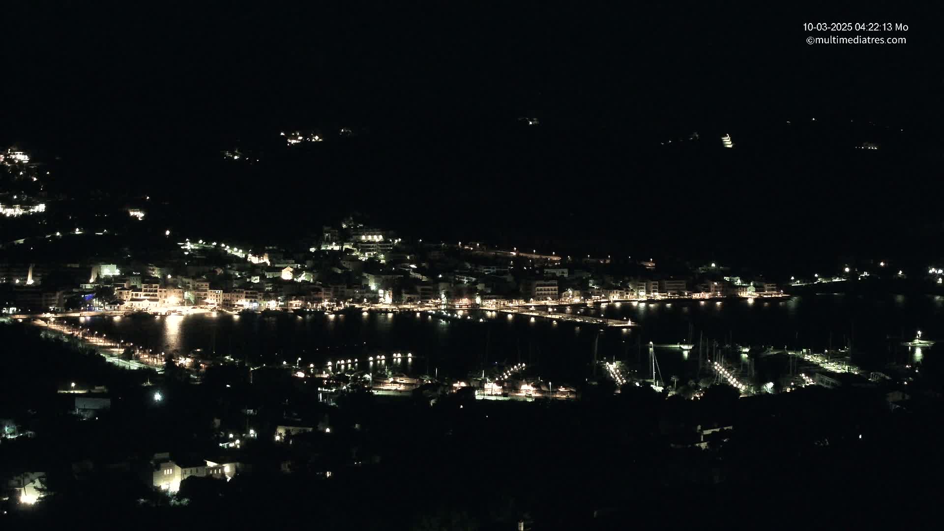 A nighttime, aerial view of a coastal town with a harbor filled with boats, all illuminated by lights.
