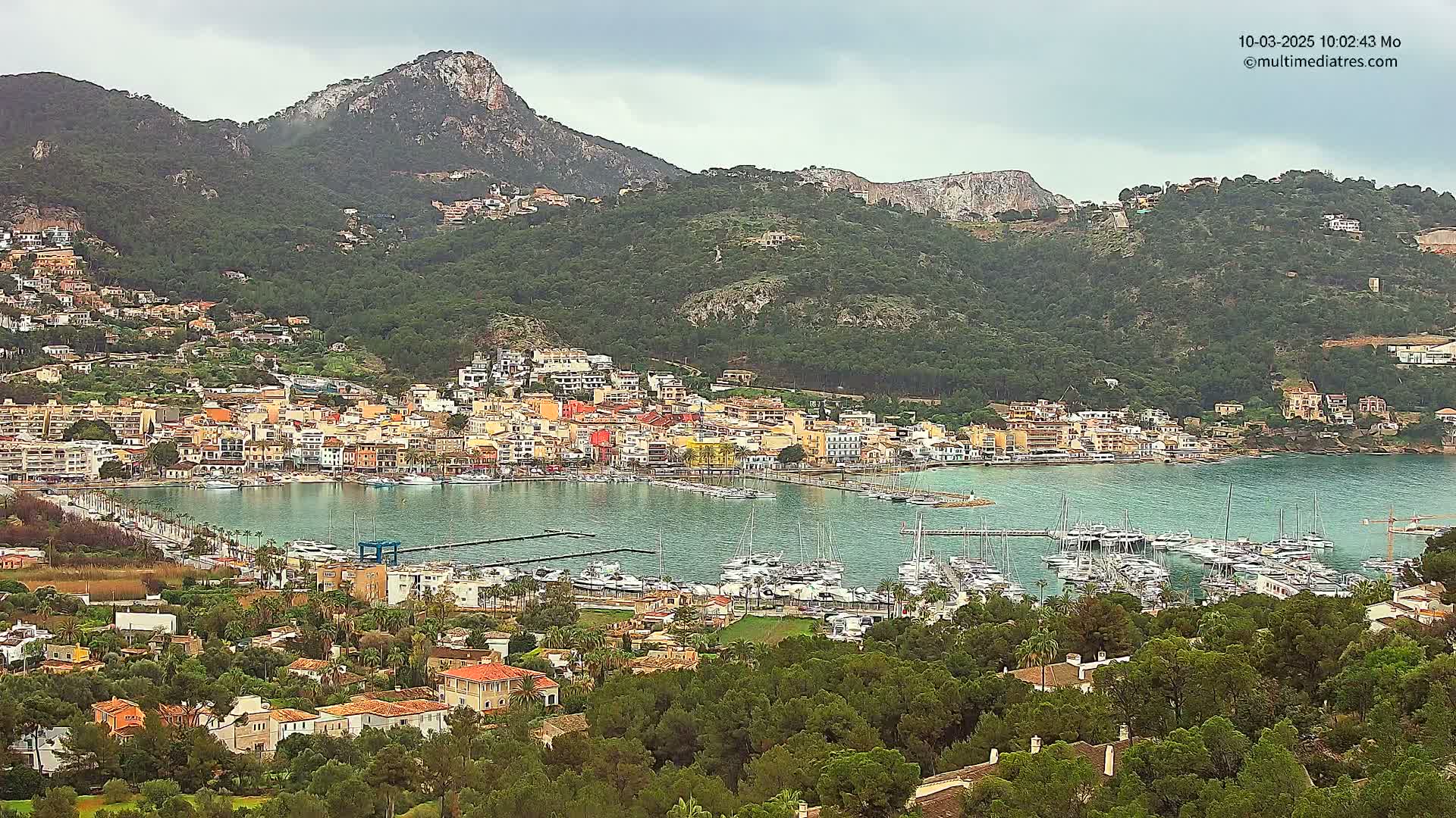 A coastal town with colorful buildings and a marina filled with boats is nestled at the foot of a green mountain range under an overcast sky.