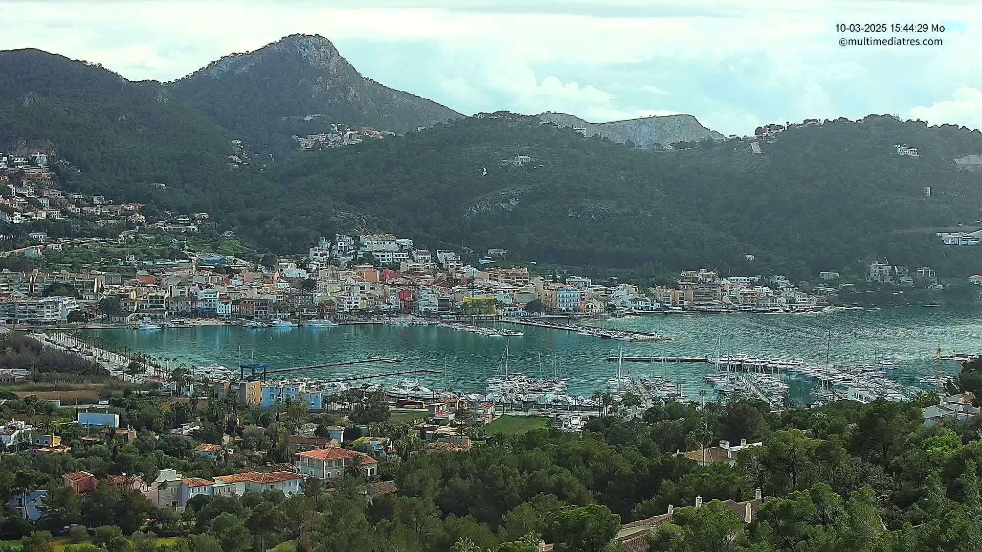 A coastal town nestled between green hills and a calm bay filled with numerous boats under a mostly cloudy sky.