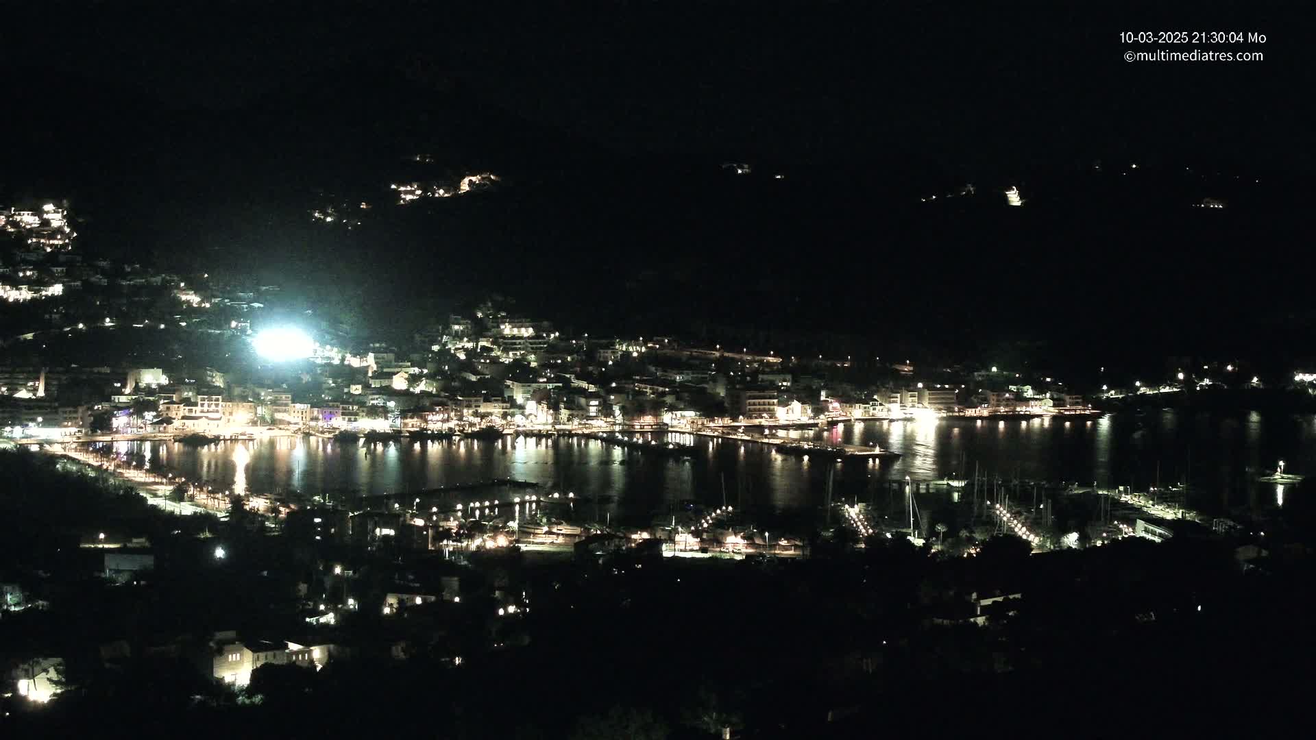 A nighttime, high-angle view shows a brightly lit coastal town nestled along a dark body of water, with many boats moored in the harbor.
