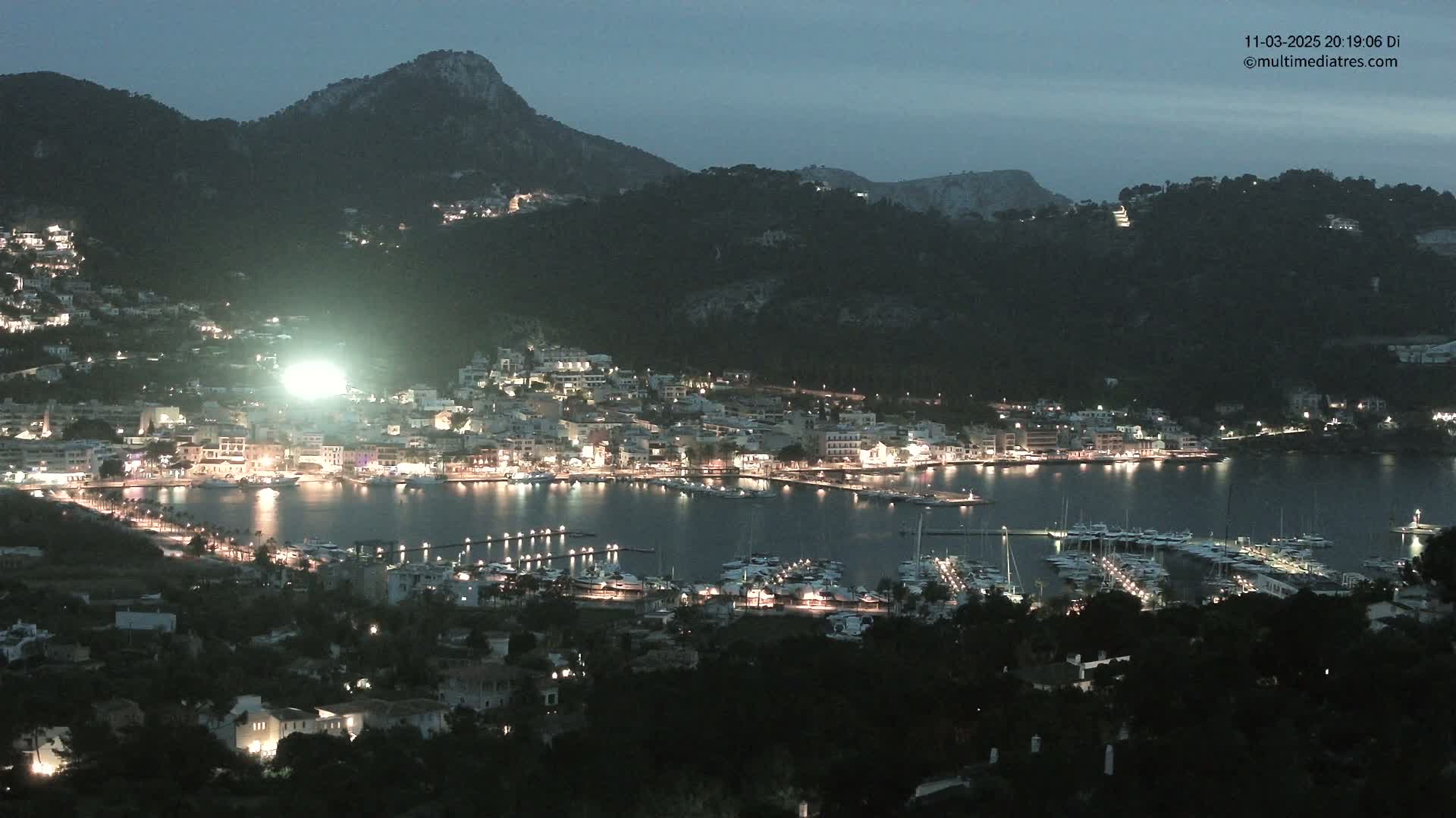 A nighttime, illuminated coastal town nestled at the base of dark, mountainous terrain with a calm harbor filled with boats.