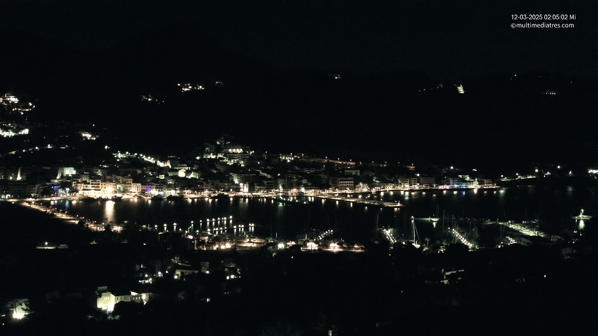 A coastal town at night, illuminated by many lights, is visible from a higher vantage point, with a calm body of water reflecting the lights.