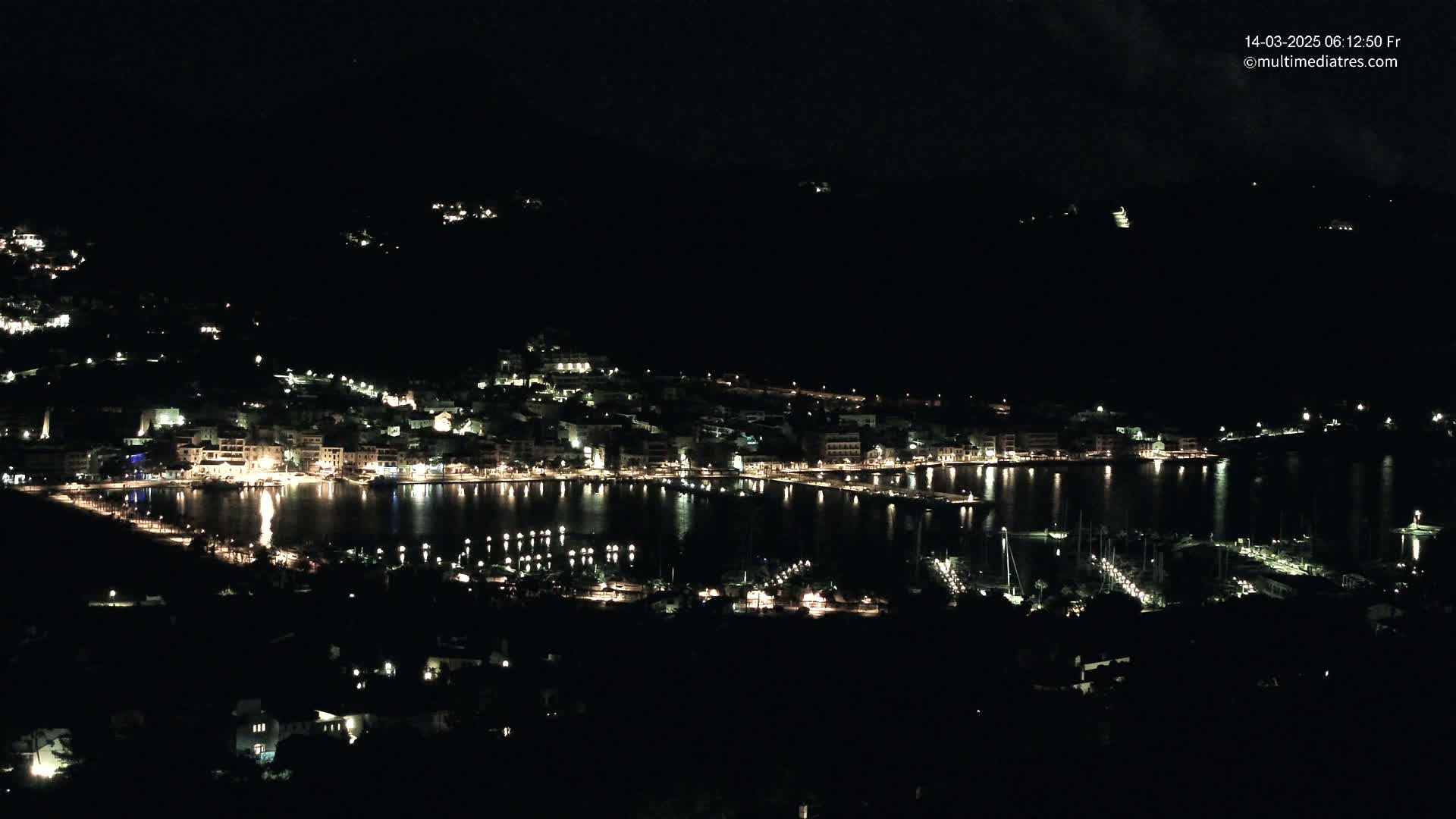 A nighttime view of a coastal town, brightly lit along its waterfront and harbor, with numerous boats moored in the calm water.