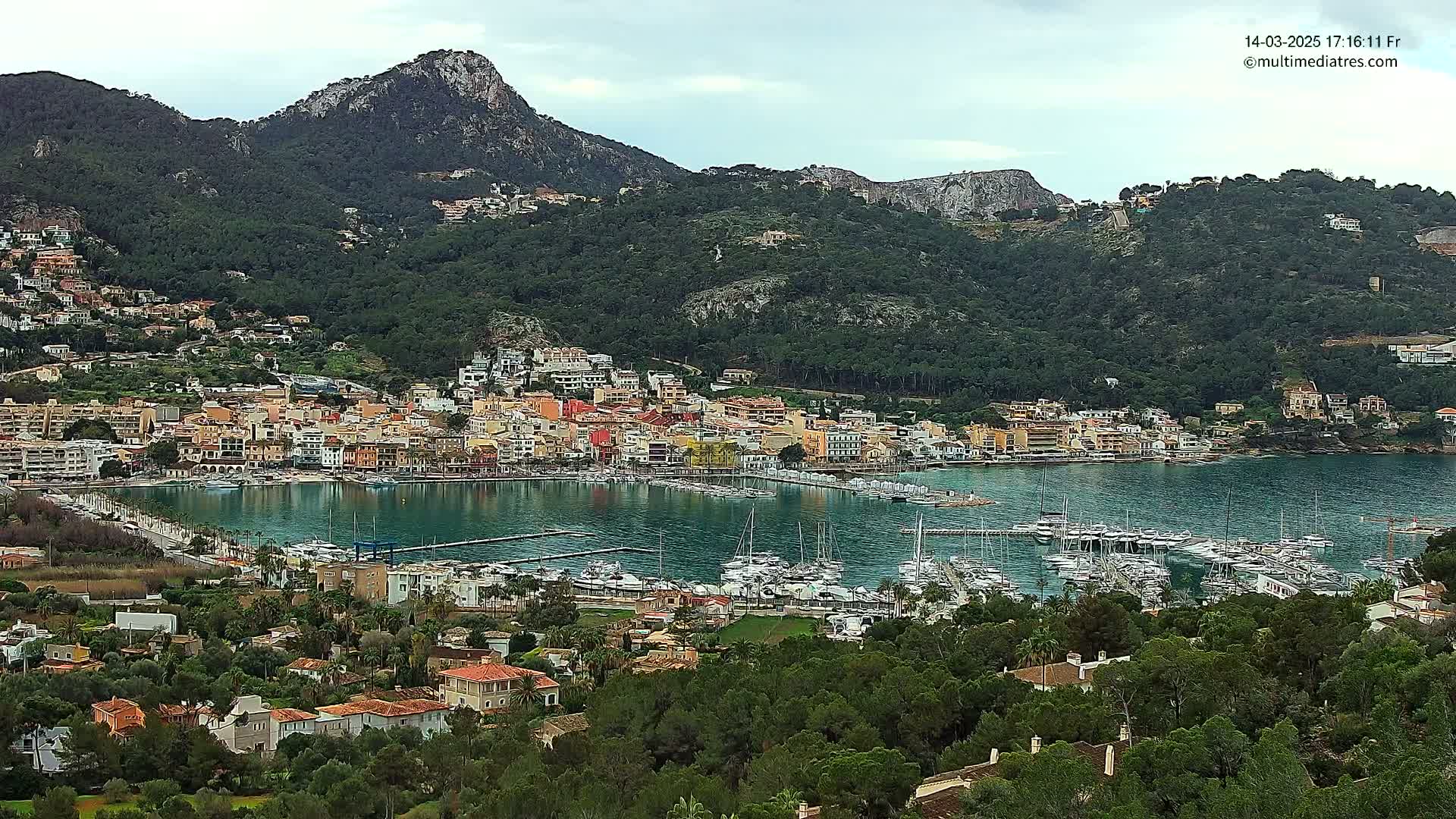 A coastal town with colorful buildings and a marina filled with boats is nestled at the base of a mountain range under an overcast sky.