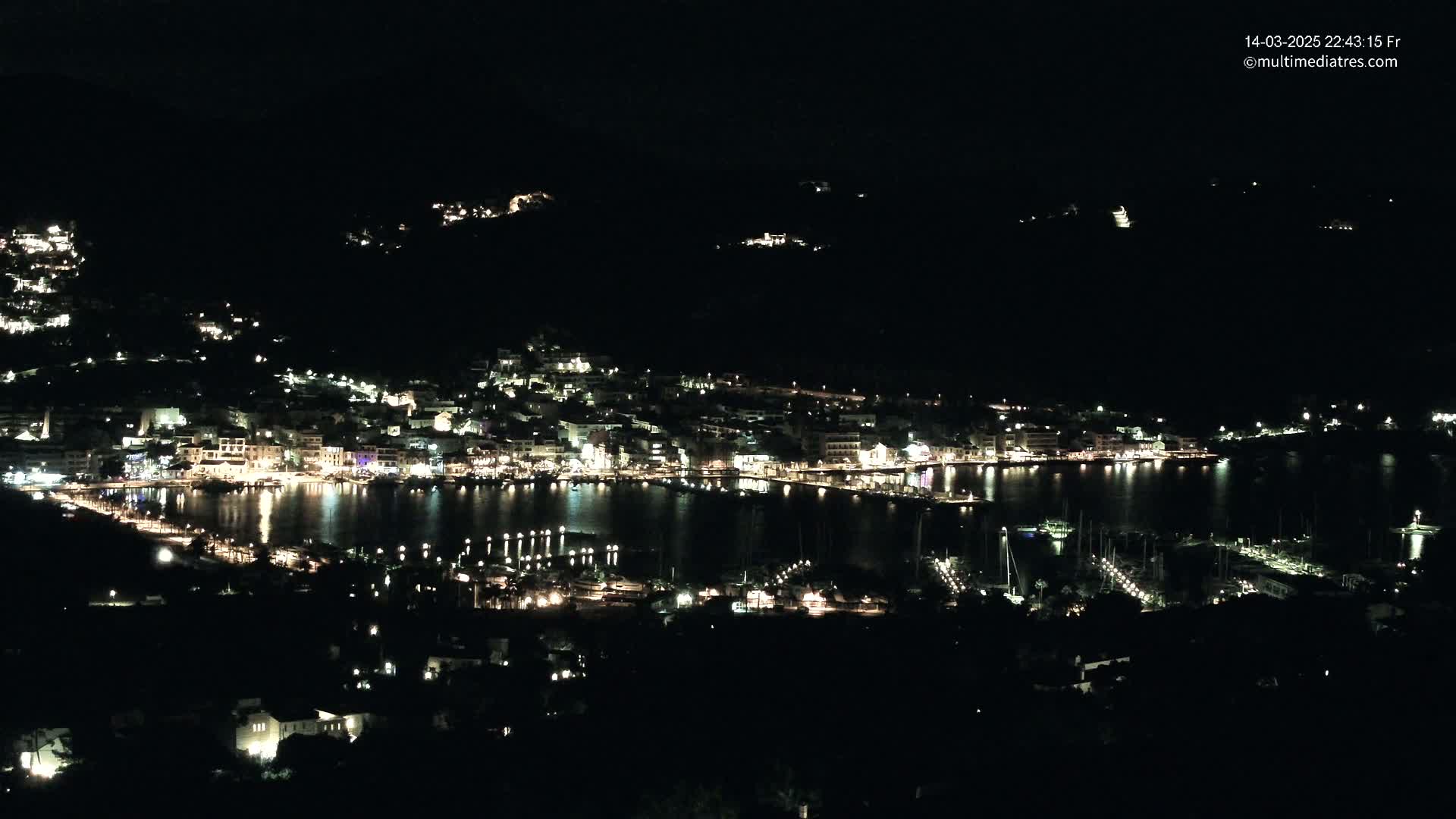 A nighttime view of a harbor town nestled on a hillside, illuminated by many lights reflecting on the calm water.