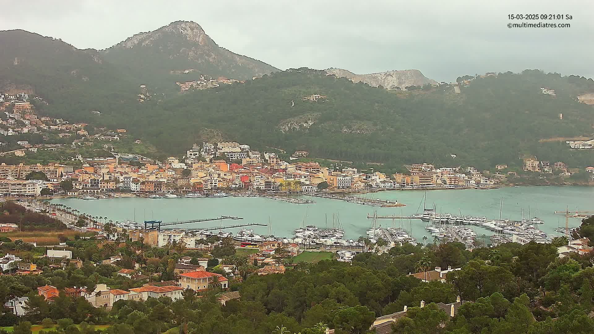 A coastal town nestled against a mountain range, with a harbor filled with boats under an overcast sky.