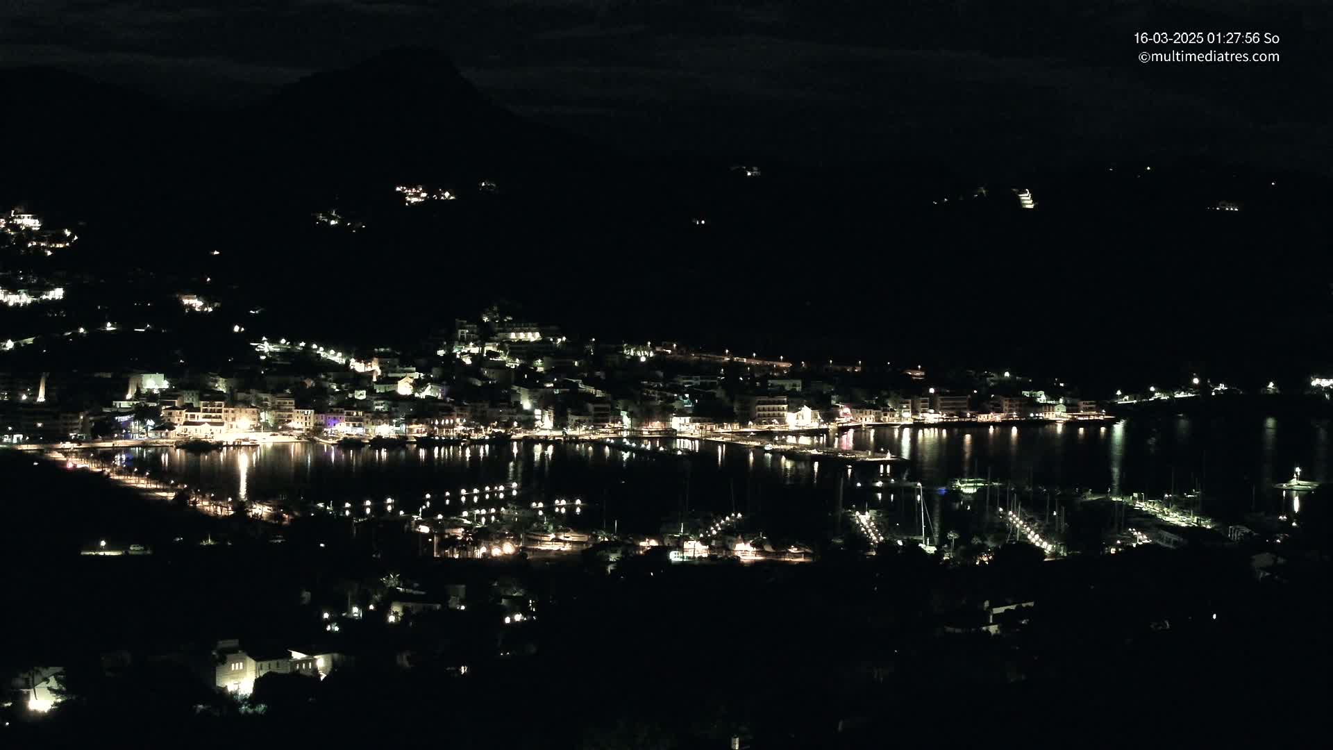 A nighttime view of a coastal town illuminated by lights, nestled against dark hills under a mostly clear, dark sky.