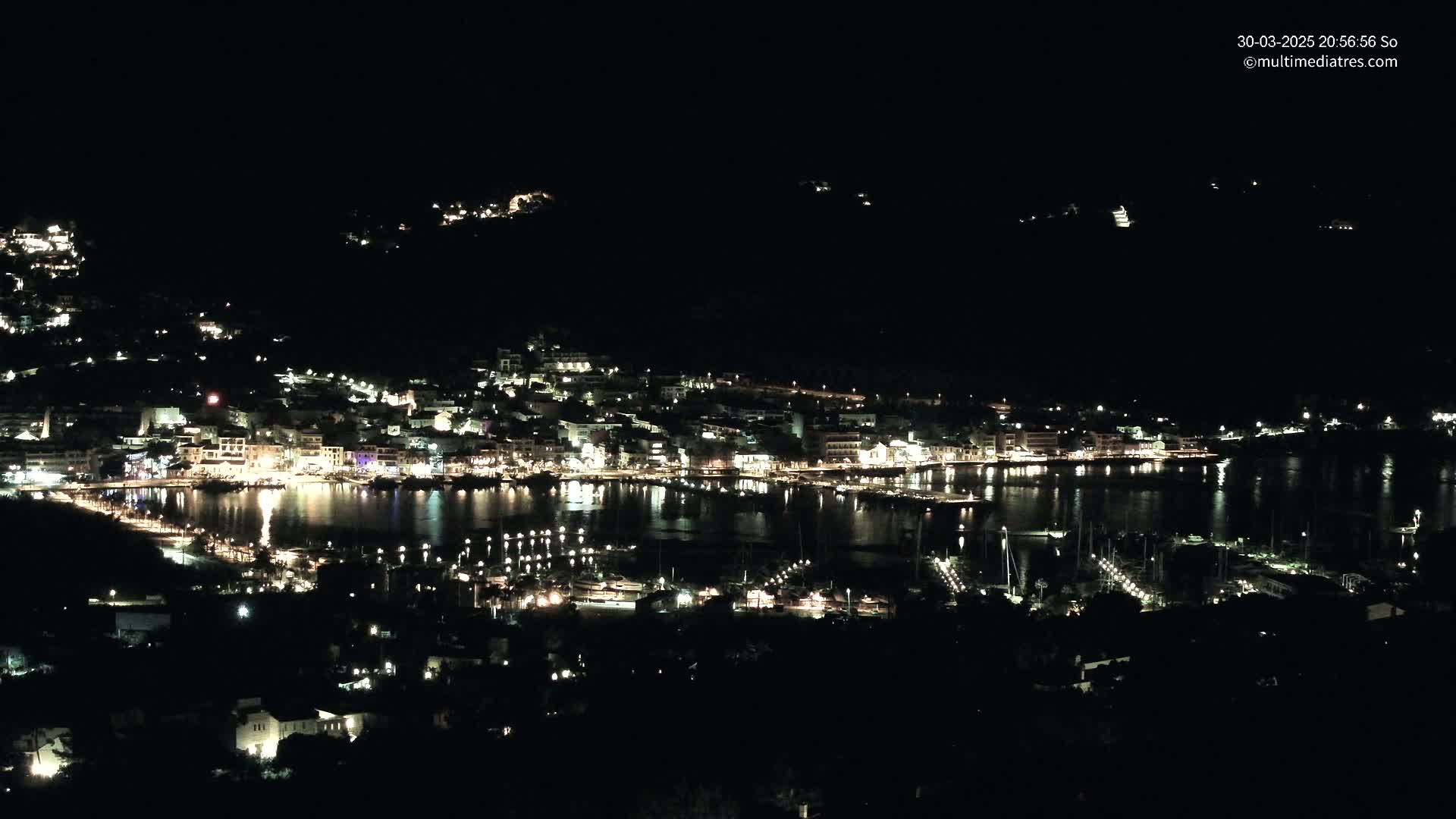 A nighttime view of a coastal town illuminated by lights, nestled between dark hills and a calm, reflective body of water dotted with boats.