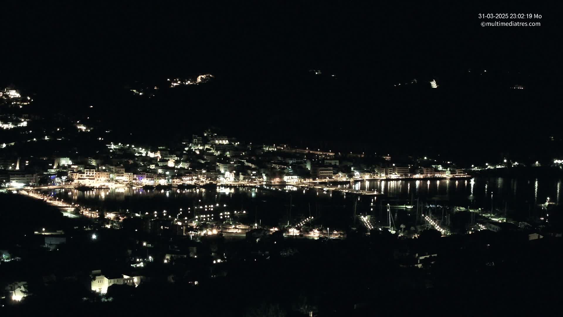 A nighttime view of a coastal town with its lights reflecting on the calm water of a harbor.
