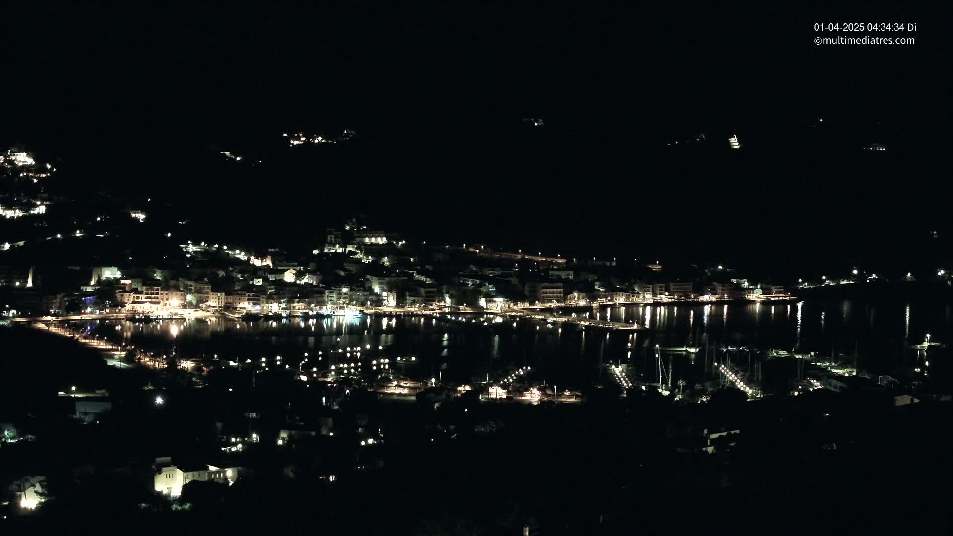 A nighttime view of a coastal town with its lights reflected in a calm body of water, surrounded by dark hills.
