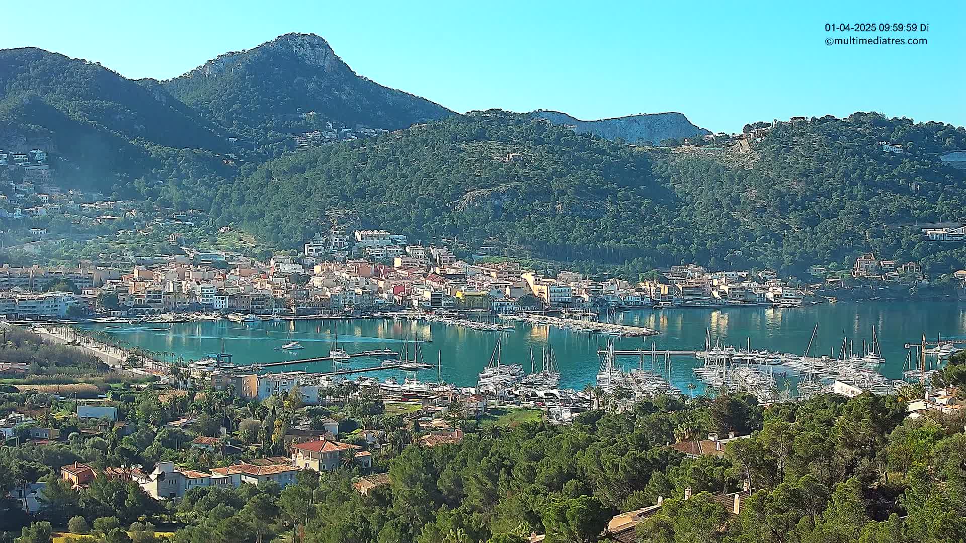 A coastal town nestled between green hills and a calm bay filled with numerous boats under a clear blue sky.