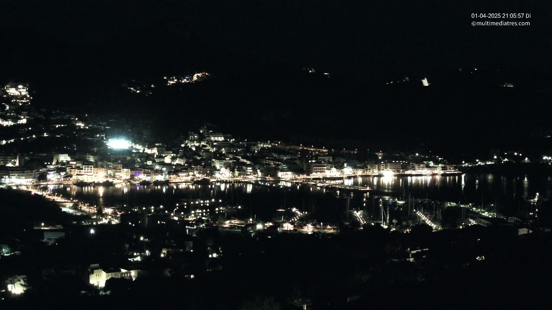 A coastal town at night, illuminated by many lights, is nestled into a dark hillside next to a calm body of water with many boats.