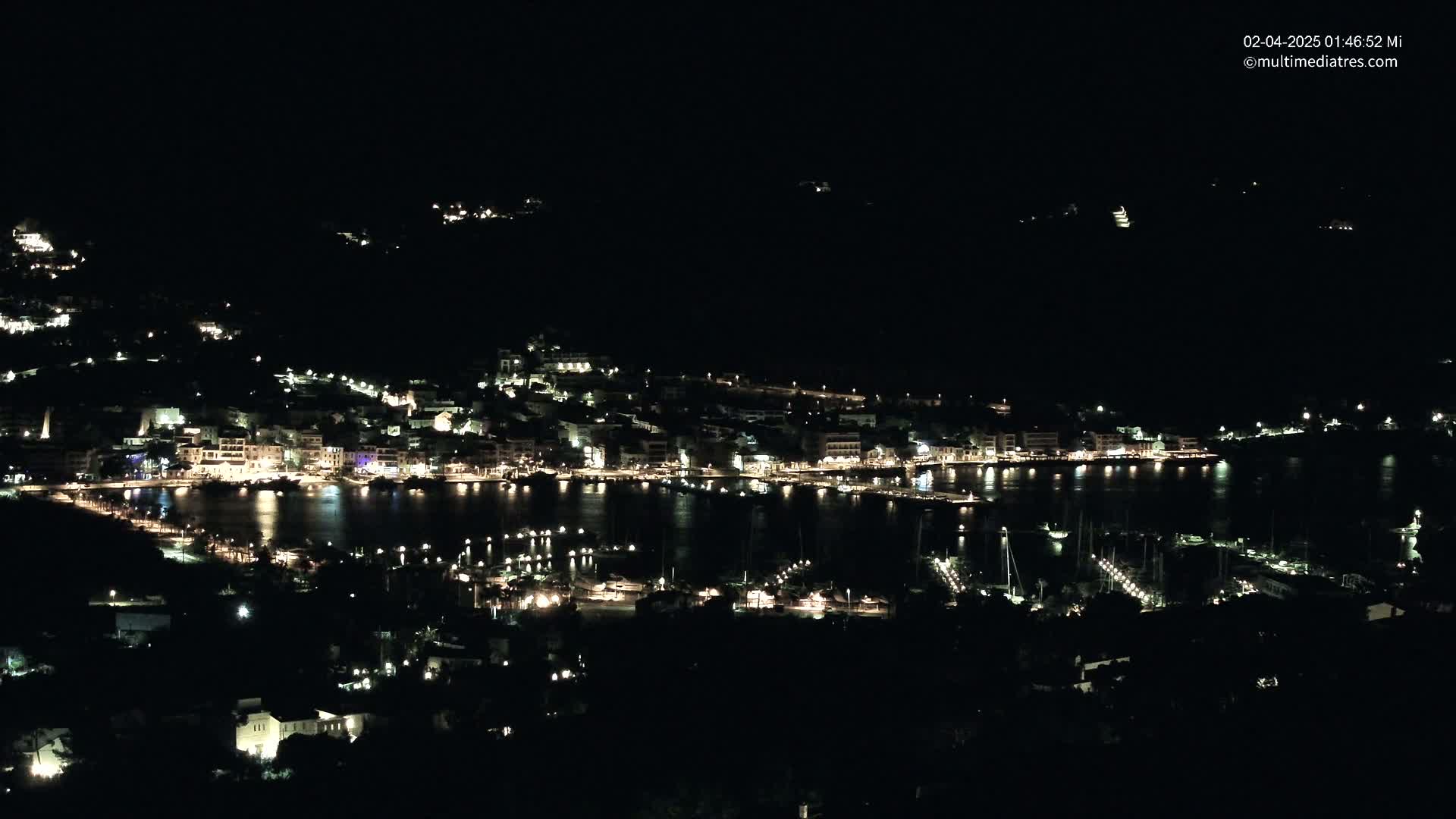 A nighttime, aerial view shows a coastal town with its lights illuminating the harbor and surrounding buildings.
