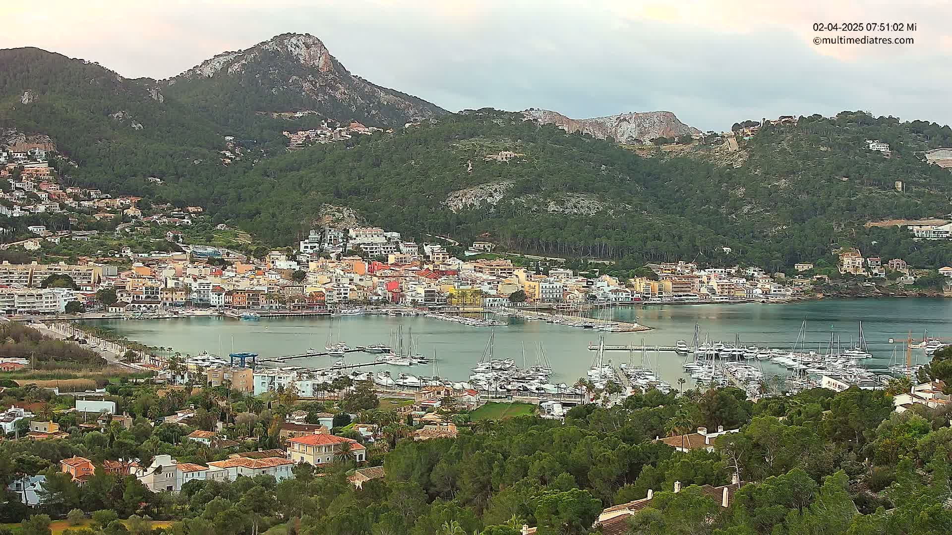 A coastal town with a marina filled with boats is nestled between green hills under a cloudy sky.