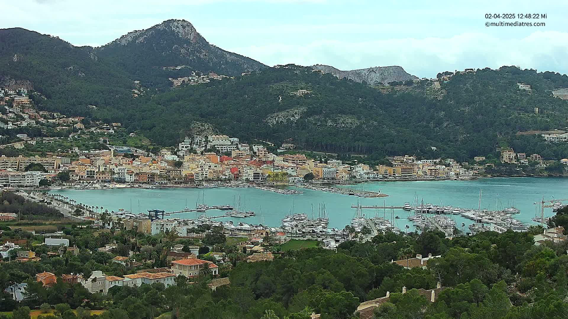A coastal town nestled between green hills and a calm bay filled with numerous boats under a cloudy sky.