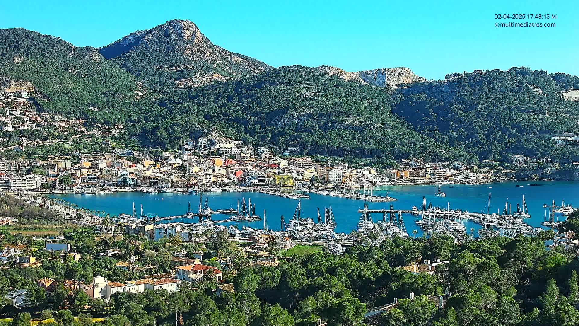 A picturesque coastal town nestled in a verdant, mountainous landscape under a clear, sunny sky, features a bustling marina filled with numerous sailboats.