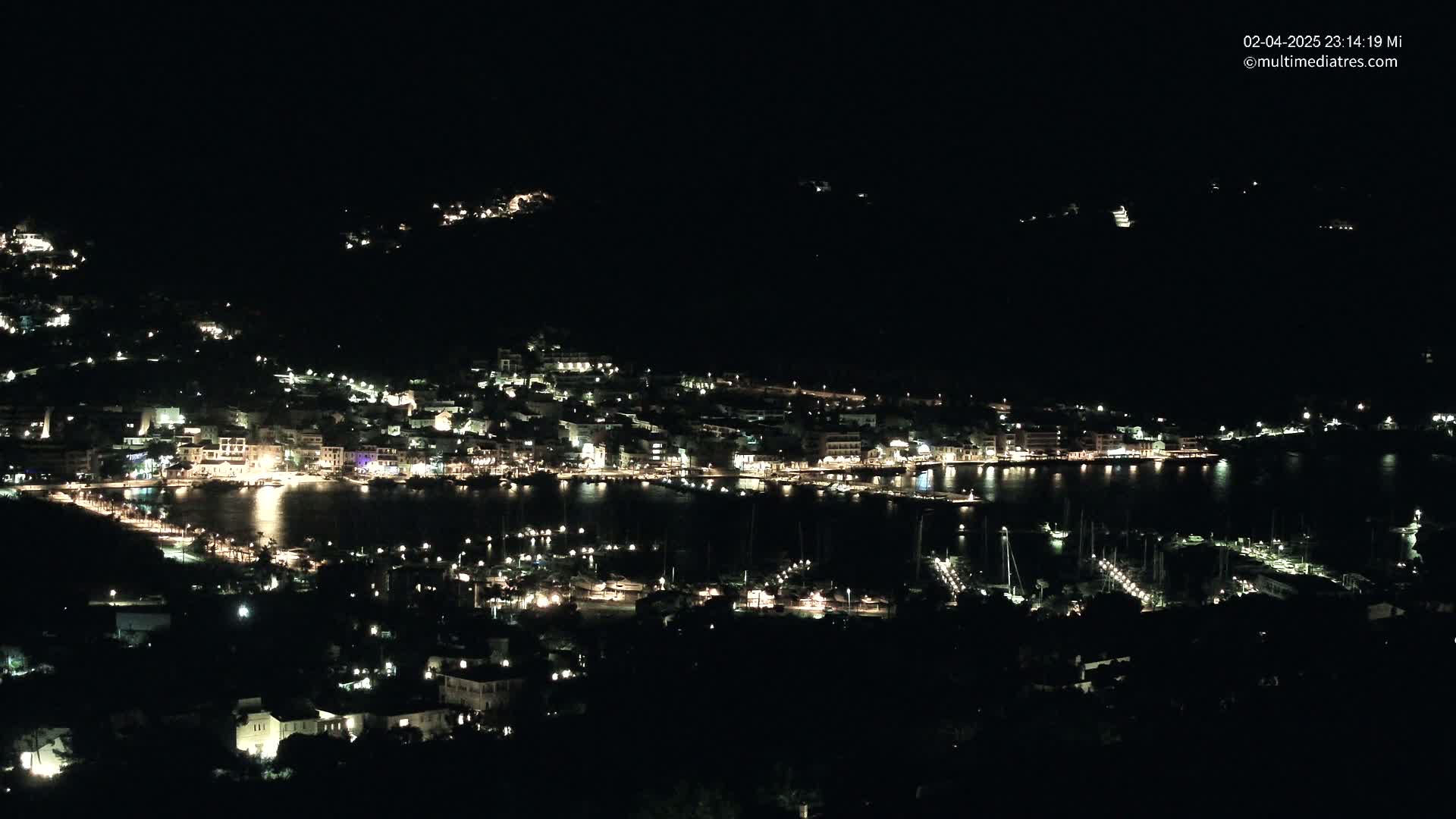 A nighttime view of a harbor town, with its buildings and a marina illuminated, nestled against dark hills under a clear, dark sky.