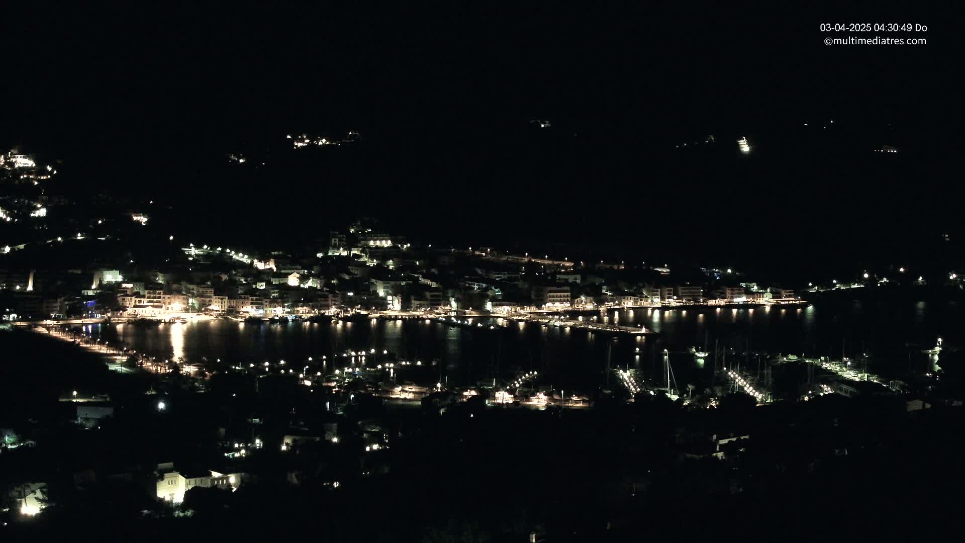 A nighttime view of a coastal town with its lights illuminating the buildings and a harbor filled with boats.