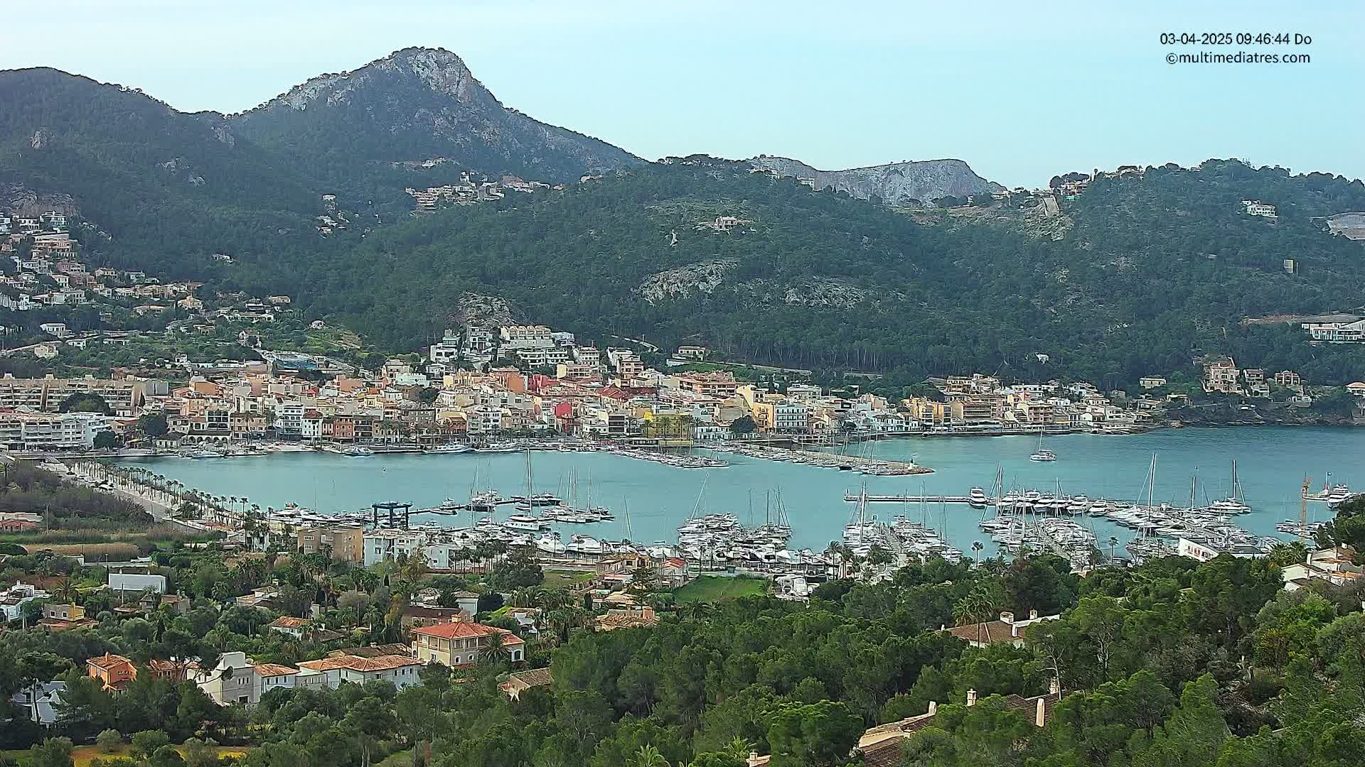 A coastal town with a marina filled with boats sits nestled at the base of a mountain range under a clear sky.