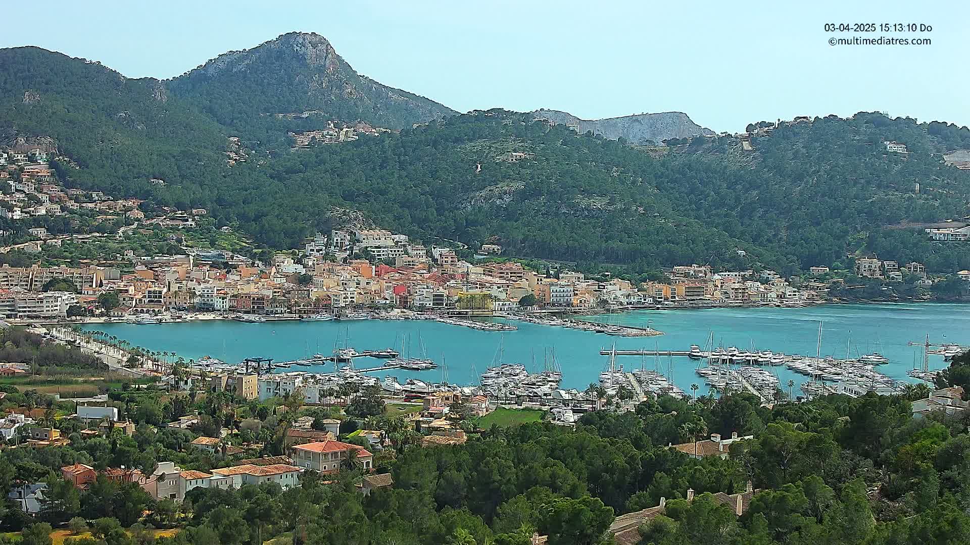 A picturesque coastal town nestled in a verdant mountain valley, with a calm harbor filled with numerous boats under a clear sky.