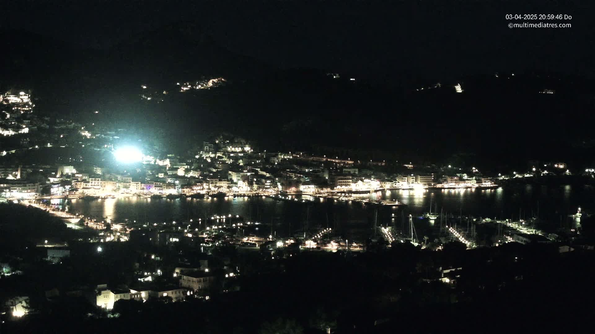 A nighttime view of a brightly lit coastal town situated on a dark hillside, with a harbor filled with boats.