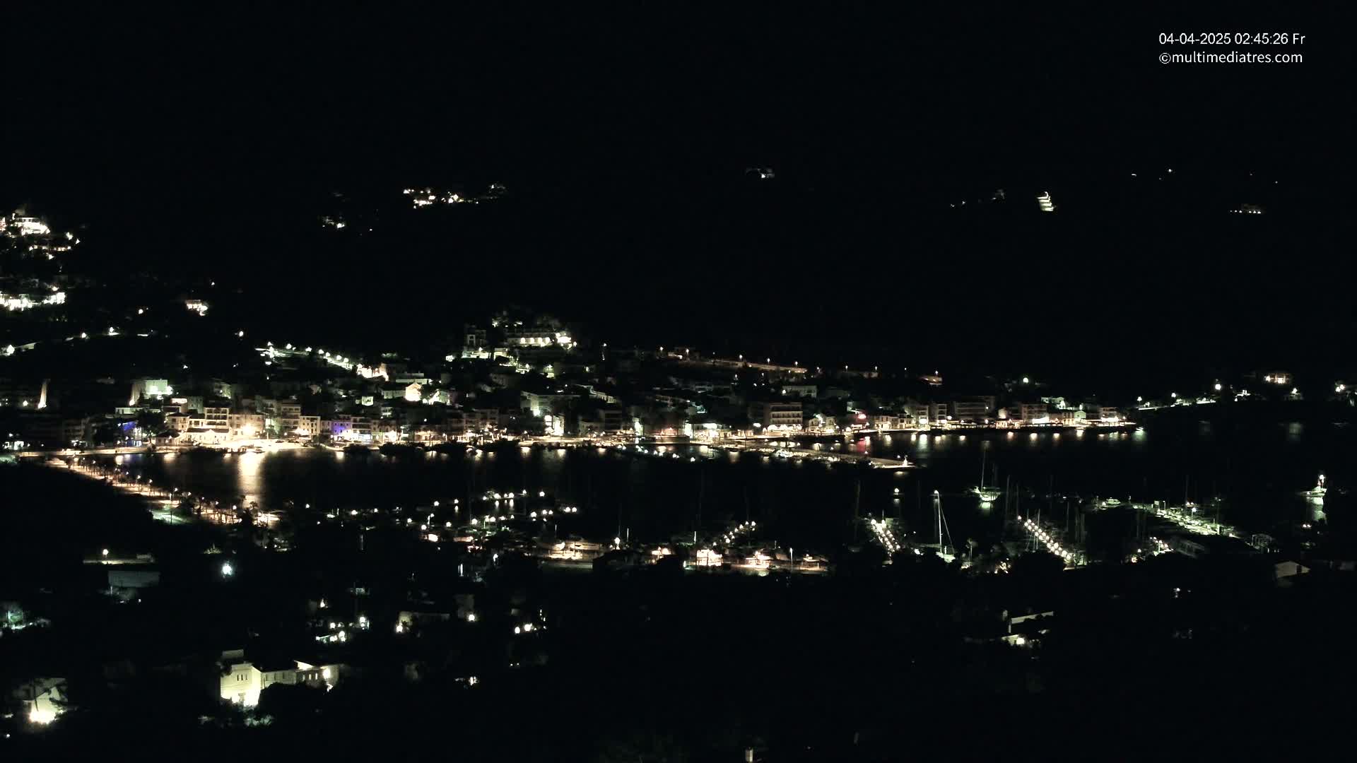 A coastal town at night, illuminated by many lights, is seen from a hilltop overlooking a harbor filled with boats.