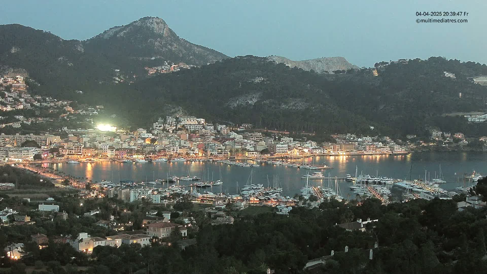 A coastal town with a marina full of boats is illuminated at twilight, nestled at the base of dark, mountainous terrain under a clear sky.
