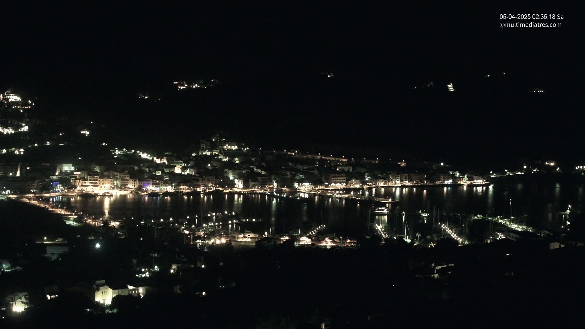 A nighttime view of a harbor town with many lights illuminating buildings and boats along the waterfront under a clear, dark sky.
