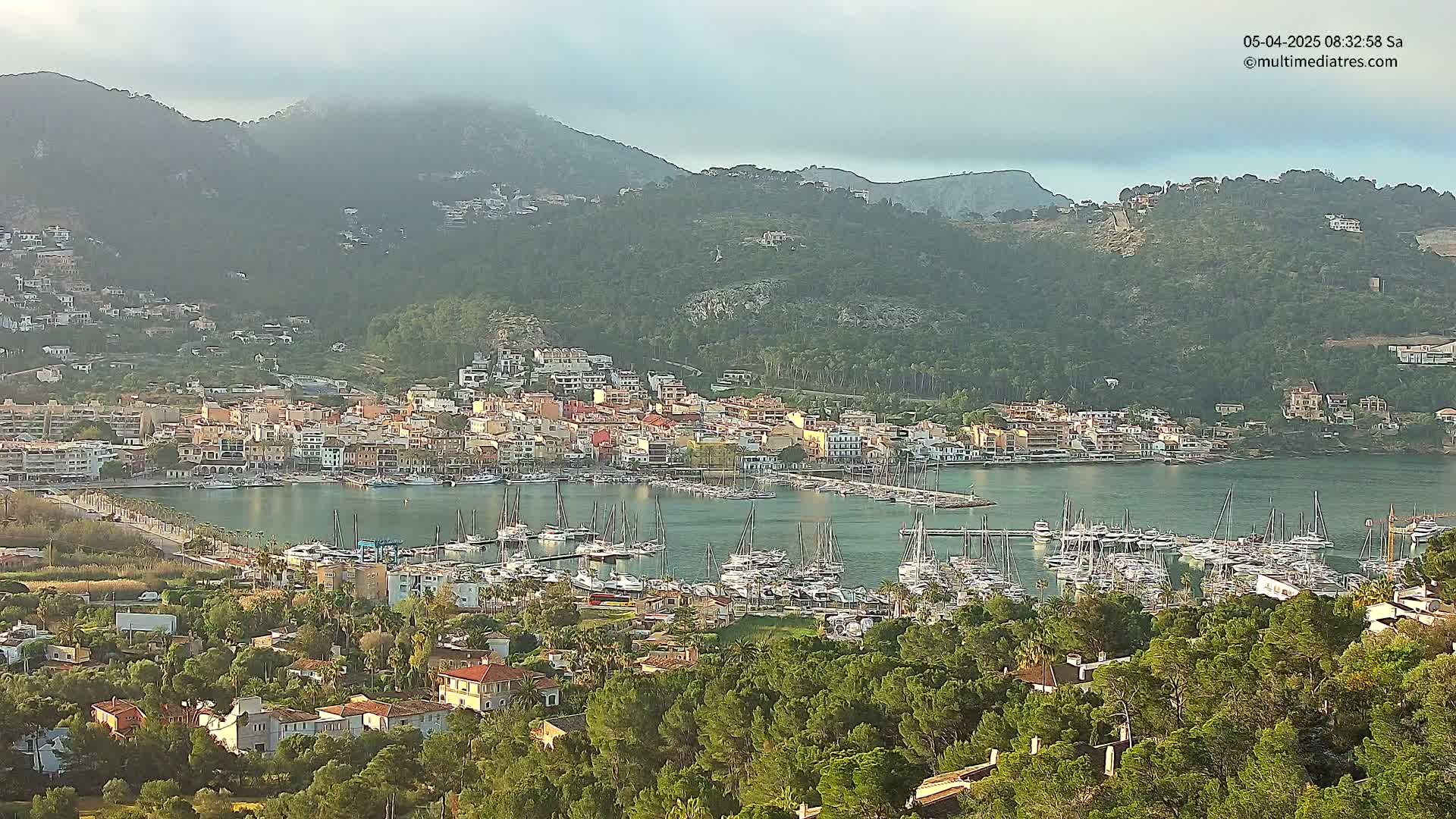 A coastal town nestled in a mountainous region with a harbor full of boats under a partly cloudy sky.