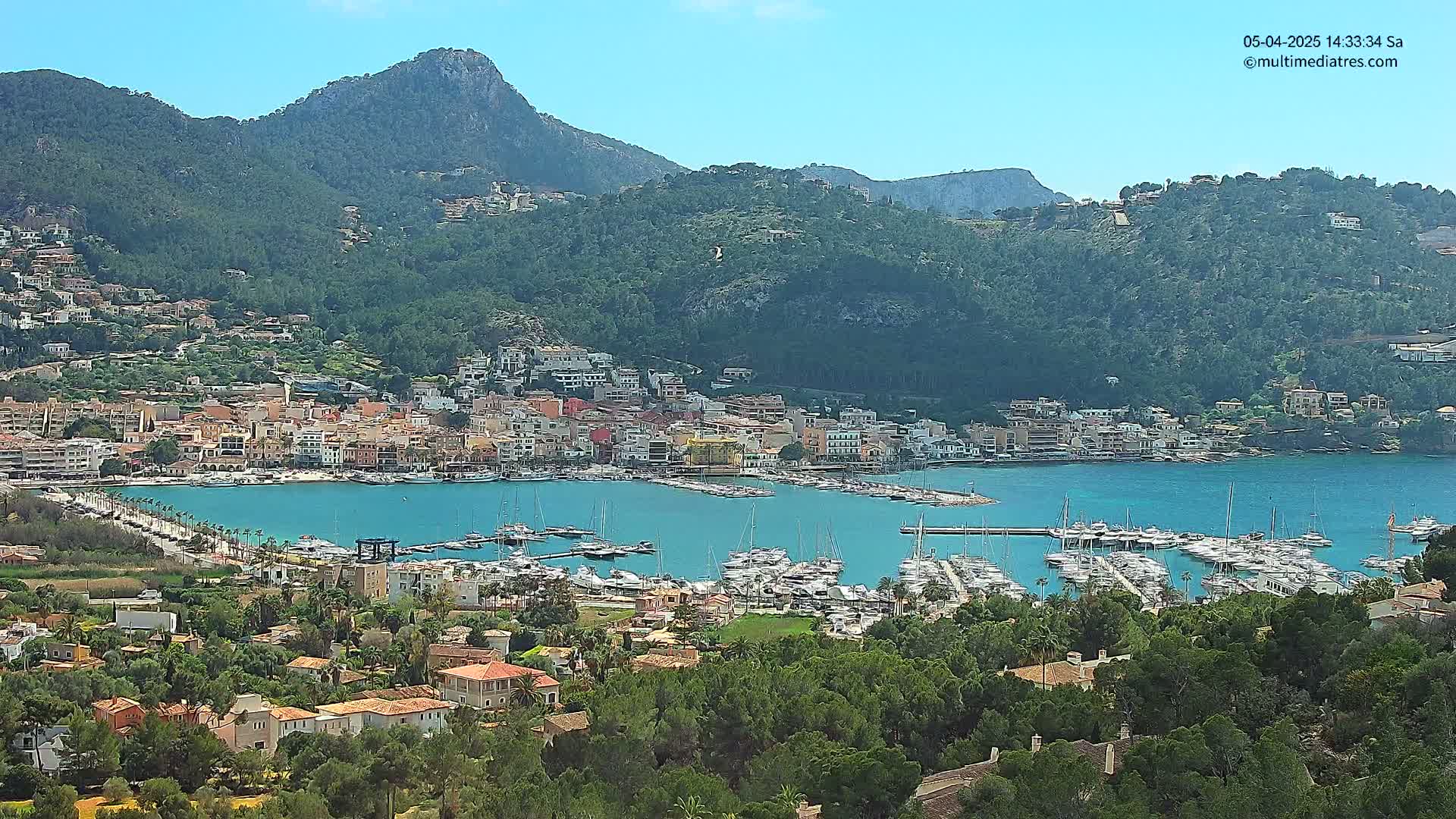 A picturesque coastal town nestled in a verdant valley, with a bustling marina filled with numerous yachts and boats, under a clear, sunny sky.