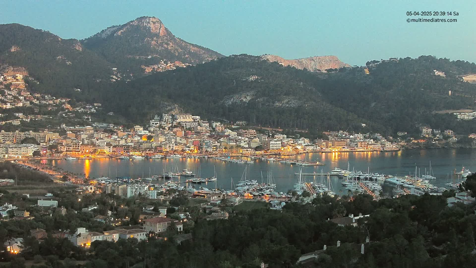 A coastal town nestled between mountains at twilight, with its harbor filled with boats illuminated by lights.