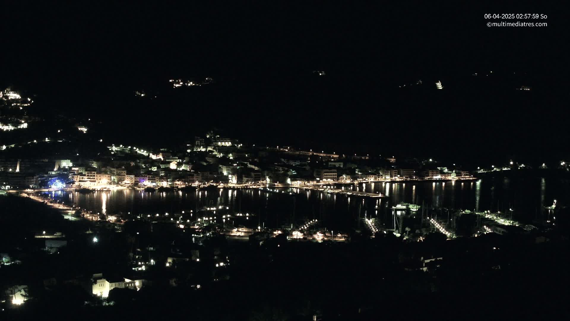 A nighttime view of a harbor town nestled on a dark hillside, with lights illuminating buildings and boats along the waterfront.