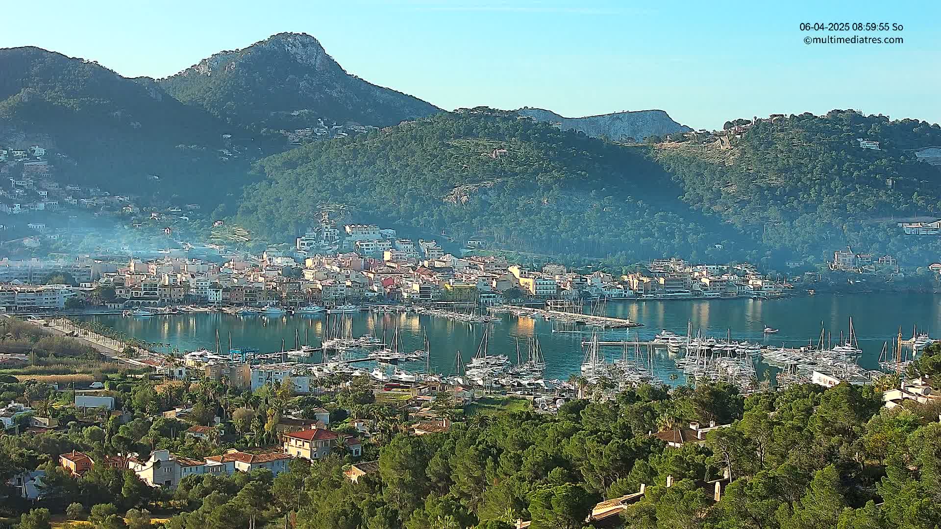 A coastal town nestled between mountains under a clear, sunny sky, with a harbor filled with numerous sailboats.