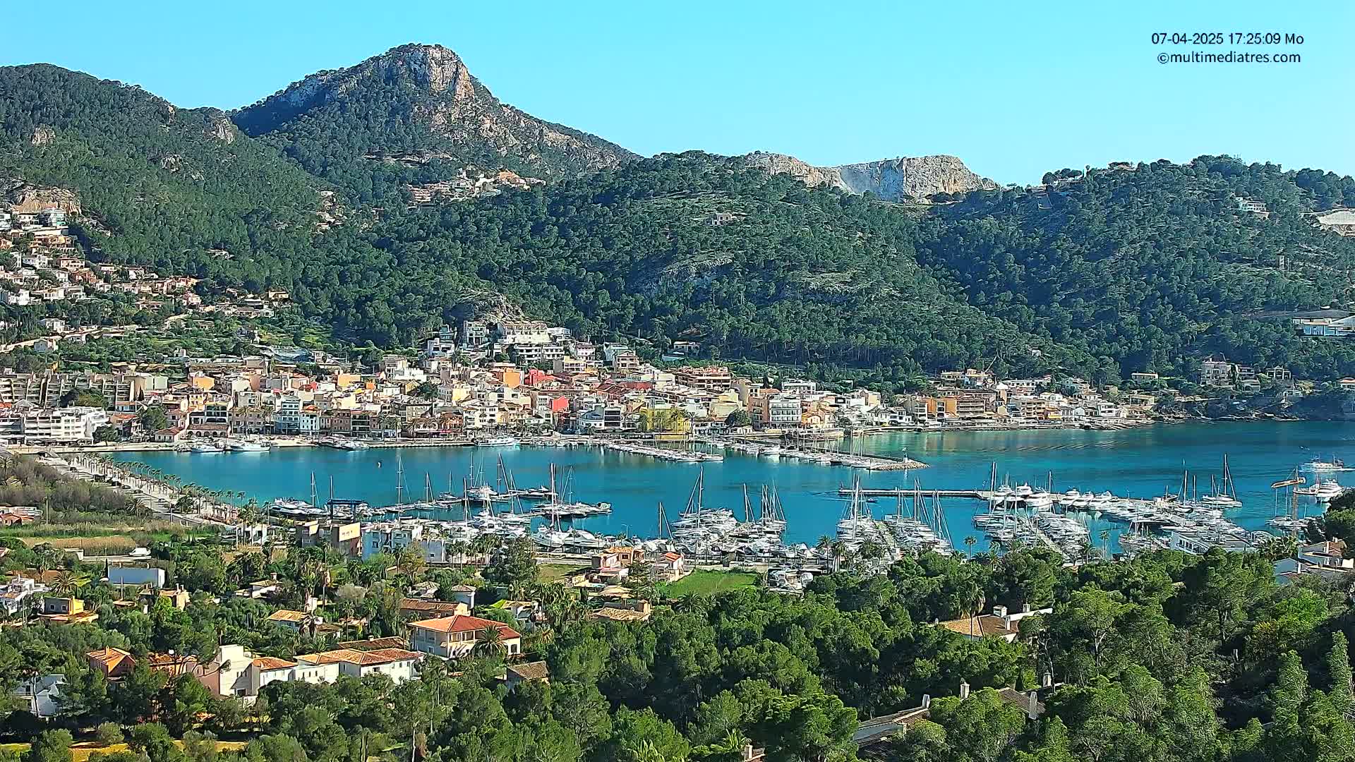 A picturesque coastal town nestled in a verdant valley, surrounded by mountains, features a bustling marina filled with numerous yachts under a clear blue sky.