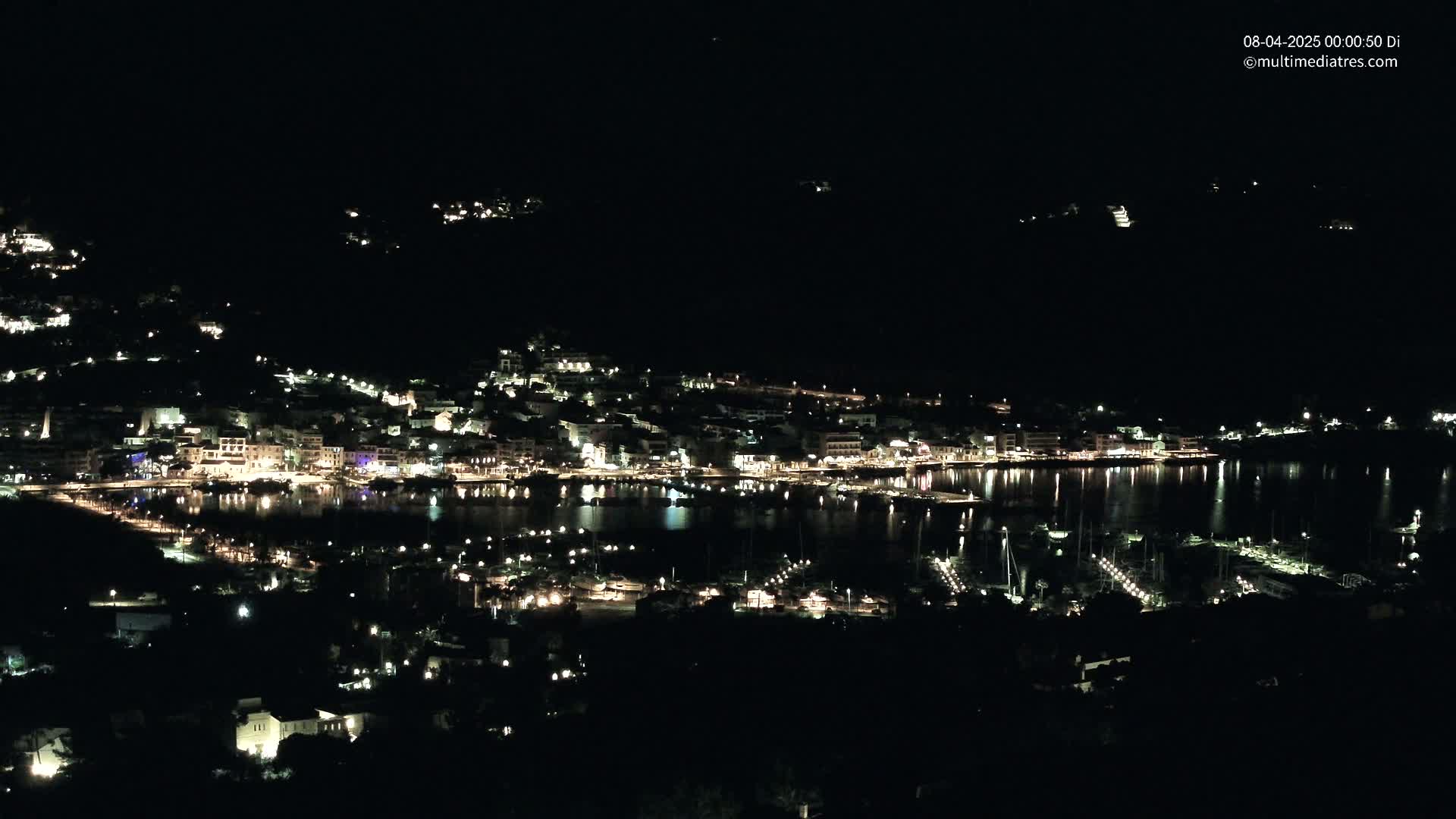 A nighttime view of a coastal town with its lights illuminating the harbor and surrounding hills under a clear, dark sky.